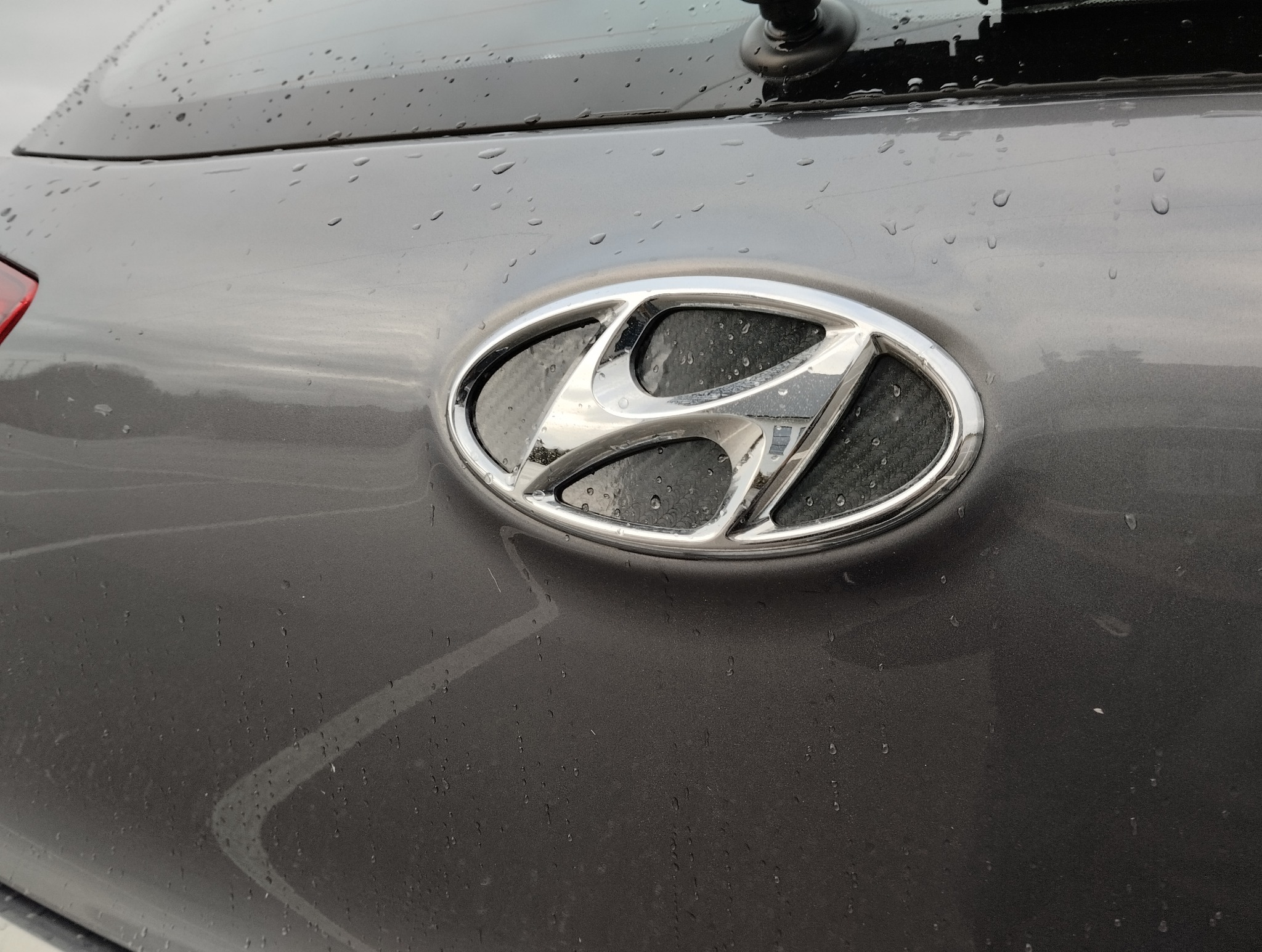 Chromowany znaczek Hyundai na mokrej, szarej karoserii samochodu. Widoczne krople deszczu i odbicia światła. Detal tylnej części pojazdu.