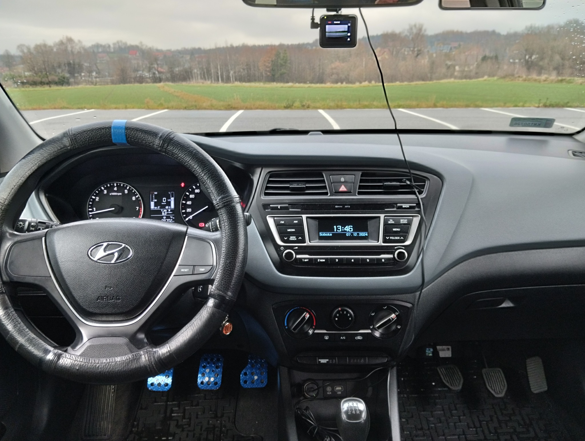 Wnętrze samochodu Hyundai i20 z widocznym kokpitem, kierownicą z niebieskim akcentem i rejestratorem jazdy. Widok na parking i drzewa za szybą.