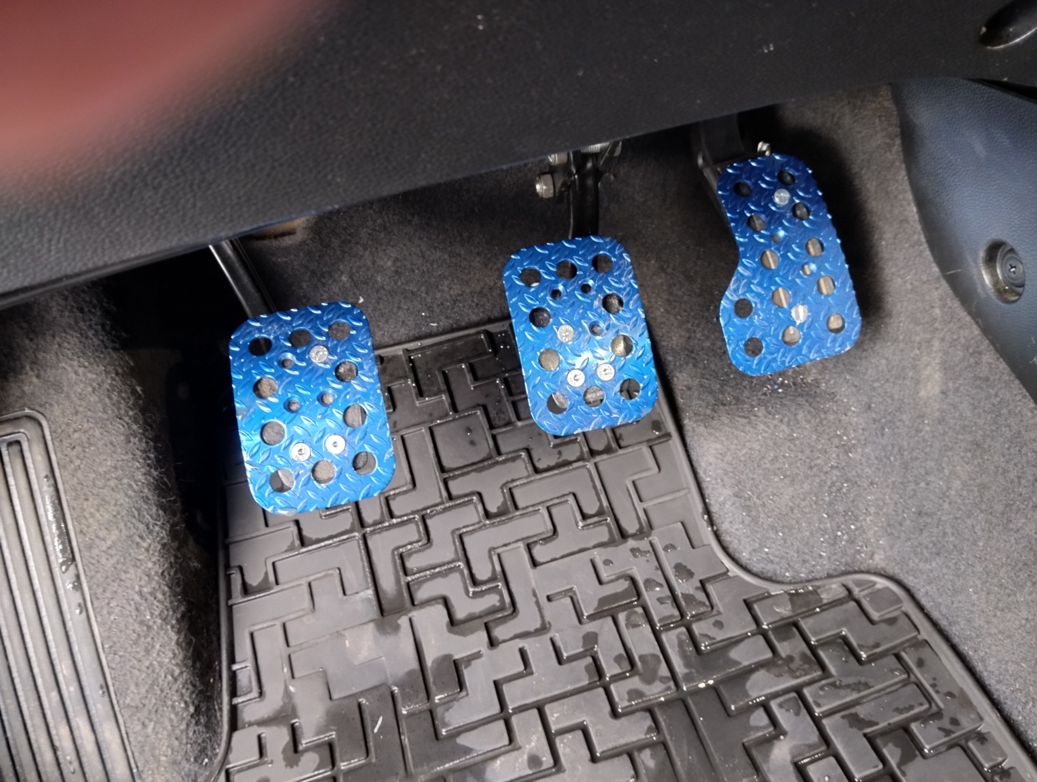 Niebieskie, metalowe nakładki na pedały w samochodzie z widoczną gumową wycieraczką podłogową. Detal wnętrza pojazdu, tuning wizualny.