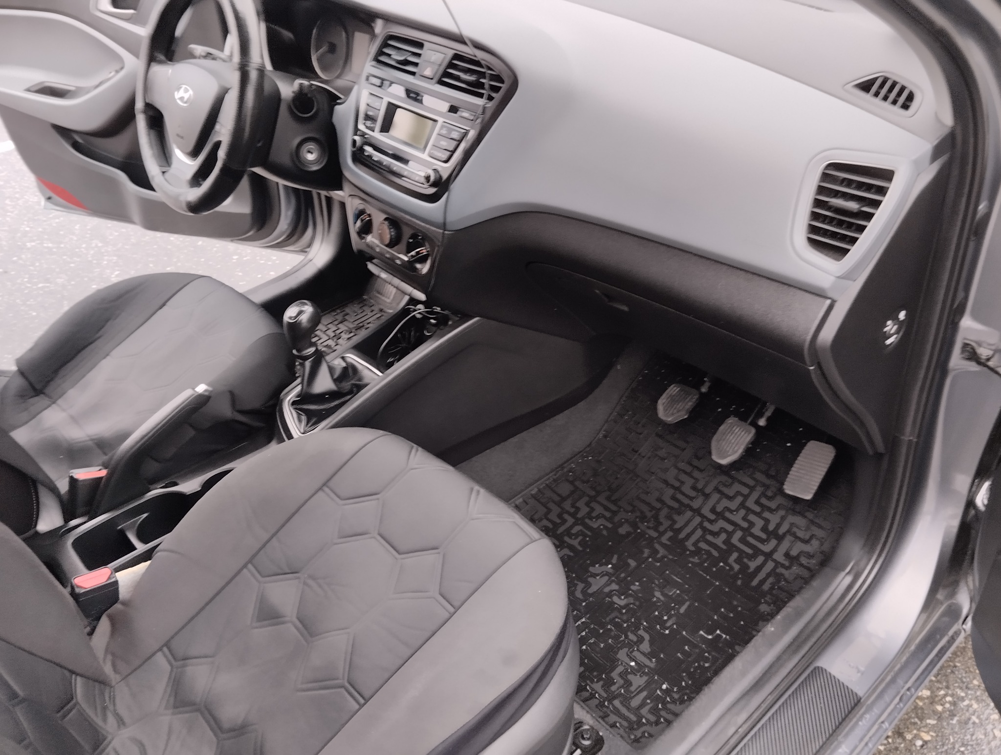 Wnętrze samochodu z widocznymi pedałami, kierownicą i deską rozdzielczą, używanego podczas kursu nauki jazdy w Łapanowie. Czarno-szare pokrowce na siedzeniach.