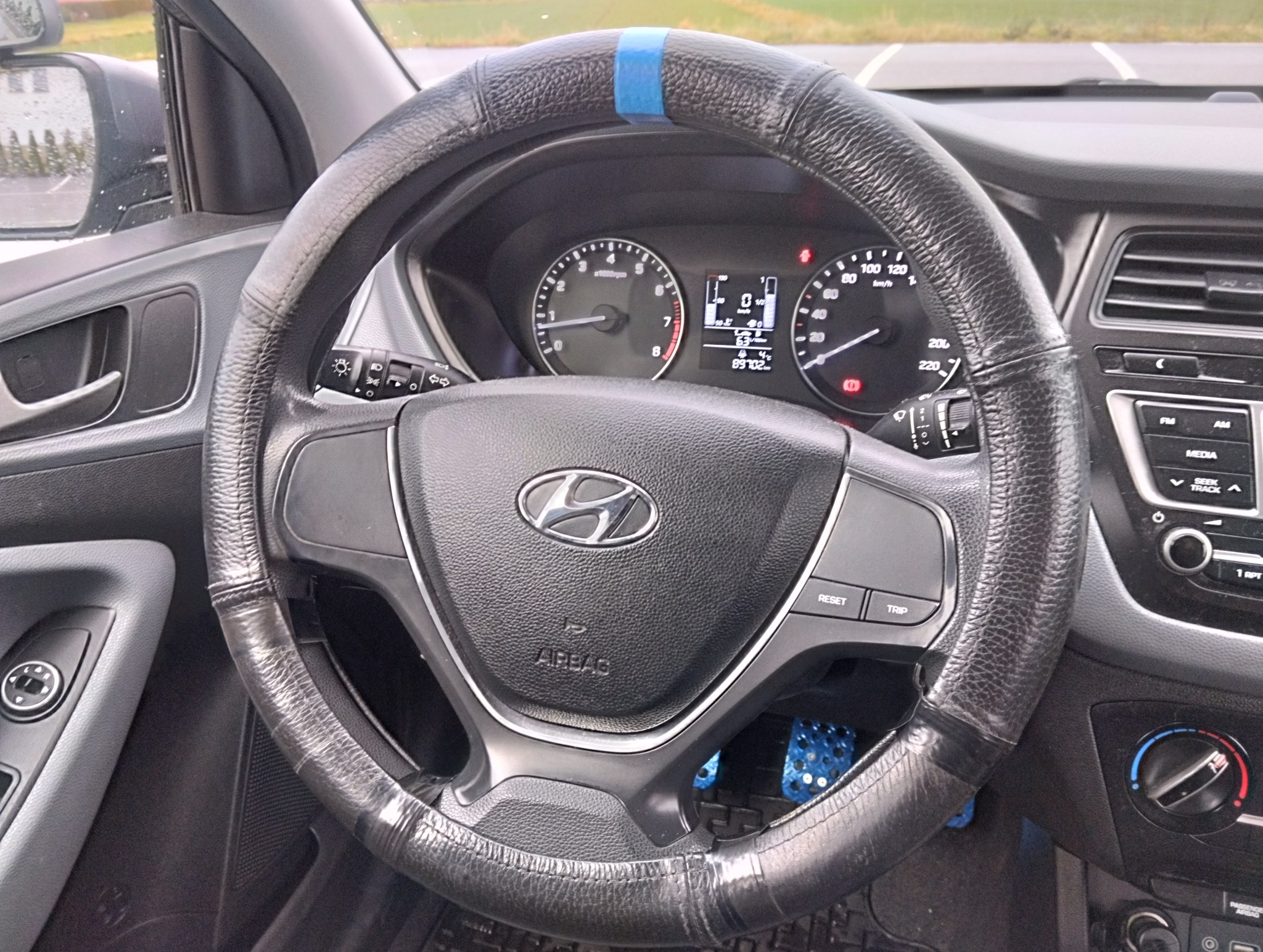 Wnętrze samochodu z widoczną kierownicą z pokrowcem i deską rozdzielczą. Na kierownicy niebieska taśma. Widoczne zegary i panel sterowania.