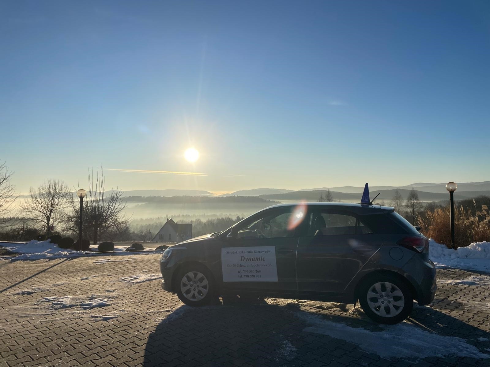 Samochód szkoły jazdy z niebieskim stożkiem na dachu, zaparkowany na tle malowniczego, mglistego krajobrazu górskiego o wschodzie słońca. Widoczny adres i telefony.