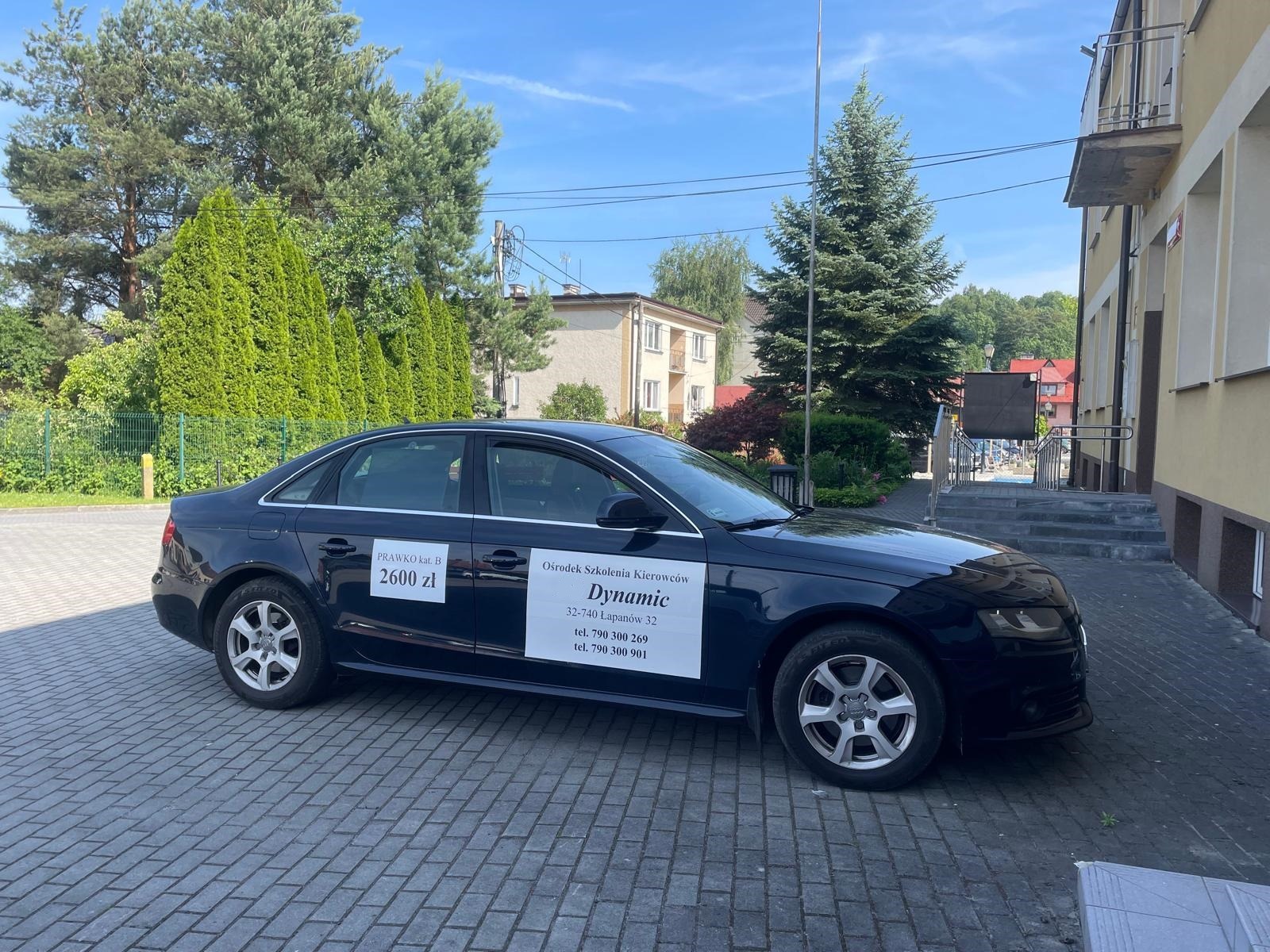 Granatowy samochód szkoły jazdy Dynamic z naklejkami informacyjnymi o cenie kursu i danych kontaktowych na tle budynku i zieleni.