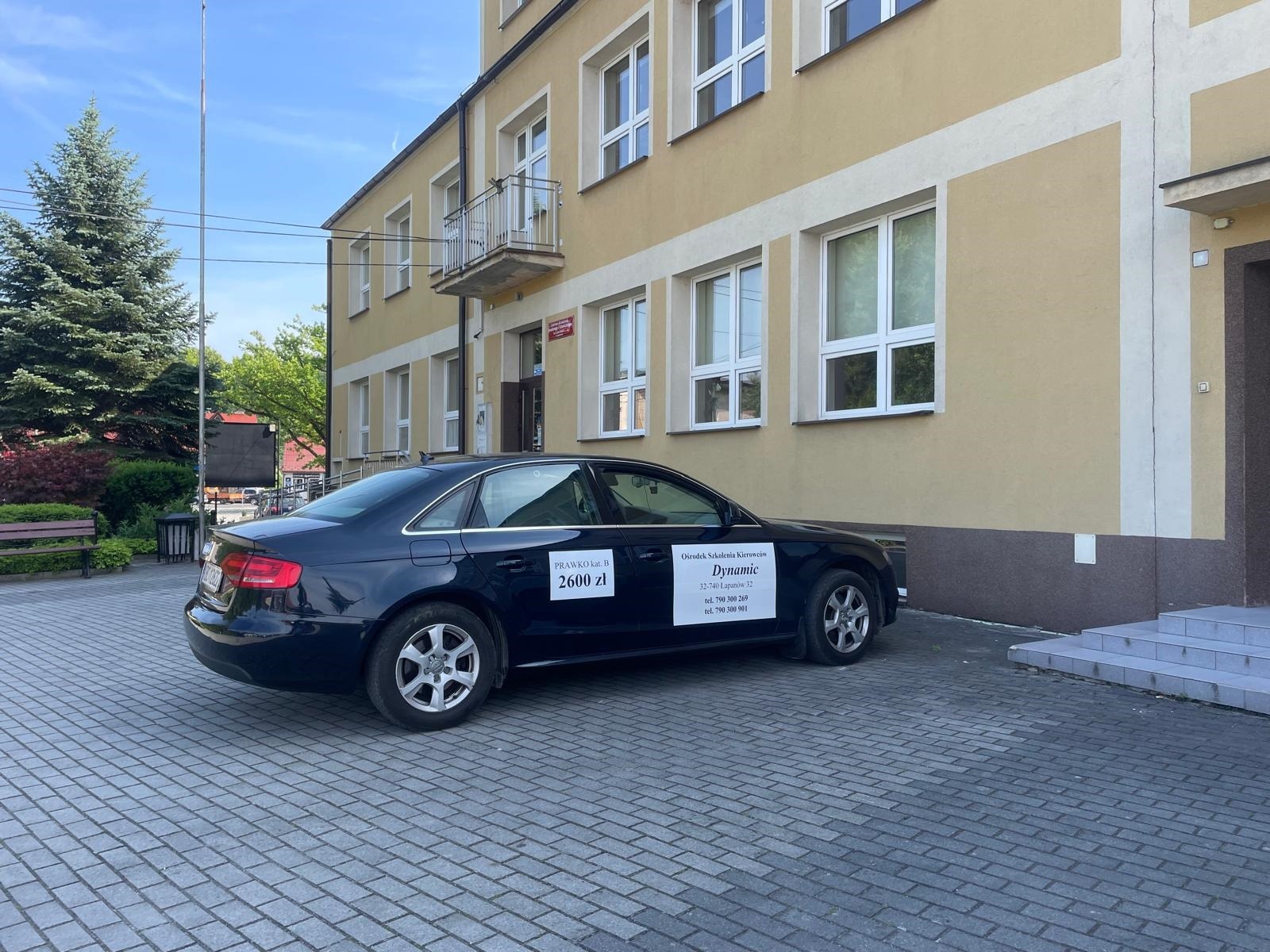 Czarny samochód z naklejkami 'PRAWKO kat. B 2600 zł' i 'Ośrodek Szkolenia Kierowców Dynamic' zaparkowany przed budynkiem szkoły jazdy.