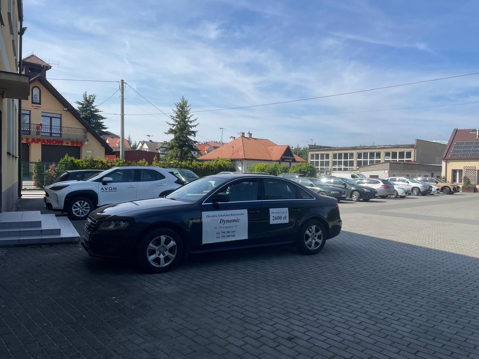 Czarny samochód z naklejką 'Ośrodek Szkolenia Kierowców Dynamic' i ceną kursu 'PRAWKO kat B 2600 zł' na parkingu przed budynkiem. W tle inne auta i budynki.