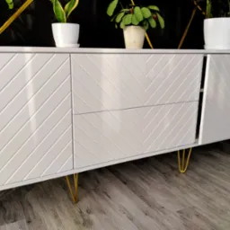 Biała komoda z frezowanym wzorem w jodełkę, na złotych nóżkach. Na blacie stoją doniczki z roślinami. Jedne drzwiczki są otwarte. Podłoga z paneli imitujących drewno.