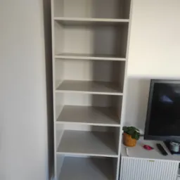 Wysoka, smukła, jasnoszara szafa regałowa z sześcioma półkami, na czarnych nóżkach, stojąca obok szafki RTV z telewizorem i dekoracjami.