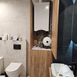 Tomasz Chmiel Hobbystycznie - Nowoczesna łazienka z zabudowanym piecem gazowym w szafce z drewnianym frontem. Umywalka nablatowa i podwieszana toaleta. Szare i czarne płytki na ścianach.