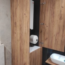 Tomasz Chmiel Hobbystycznie - Szafka łazienkowa z drewnianymi frontami, częściowo otwarta, ukrywająca bojler. Widoczna umywalka nablatowa i czarne płytki w tle. Ujęcie z bliska.