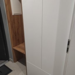 Tomasz Chmiel Hobbystycznie - Biała szafa wnękowa z drewnianym akcentem i siedziskiem, widoczna przy wejściu do mieszkania. Minimalistyczny design, jasne kolory, nowoczesne wykończenie.