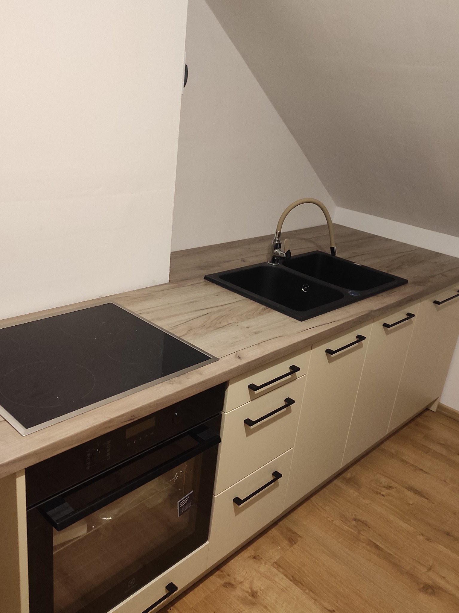 Kremowa zabudowa kuchenna pod skosem z drewnianym blatem, czarnym zlewem i płytą indukcyjną. Uwagę zwracają czarne, podłużne uchwyty szafek i szuflad.