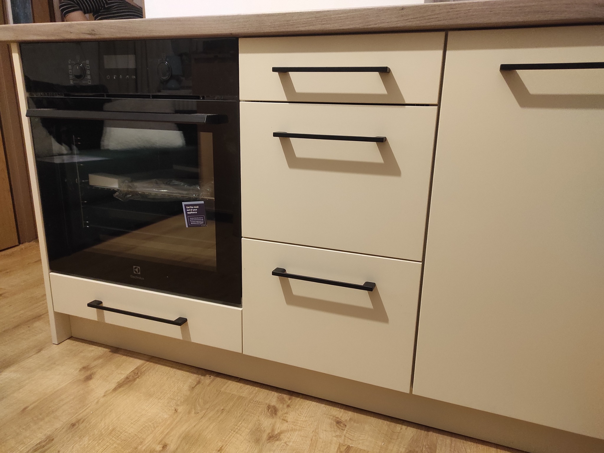 Beżowa zabudowa kuchenna z piekarnikiem Electrolux i szufladami z czarnymi, prostokątnymi uchwytami. Blat w kolorze jasnego drewna. Widoczna faktura paneli podłogowych.