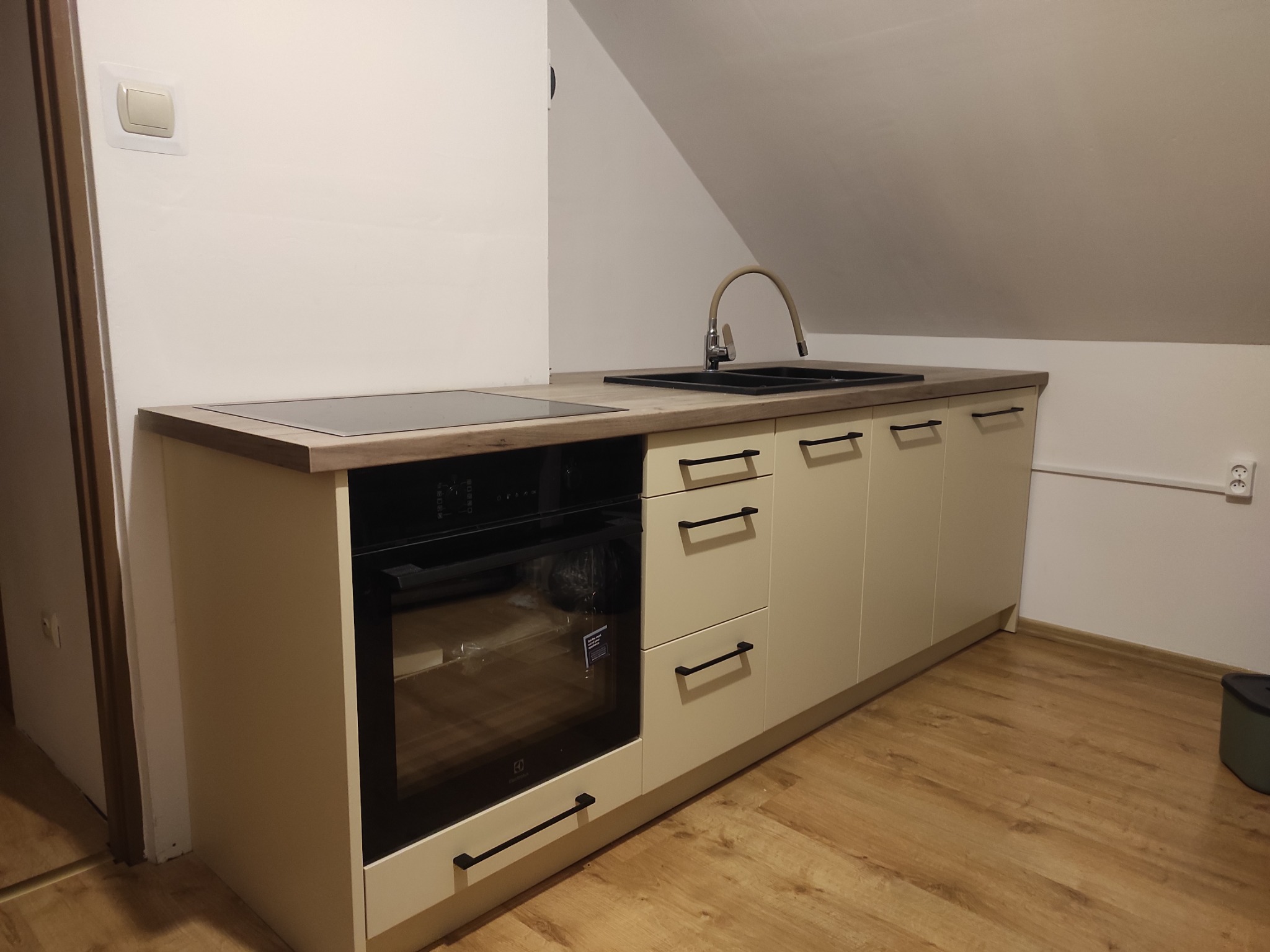 Nowoczesna kuchnia w odcieniach beżu z czarnymi uchwytami, wkomponowana w przestrzeń pod skosem. Widoczny piekarnik Electrolux i drewniany blat.