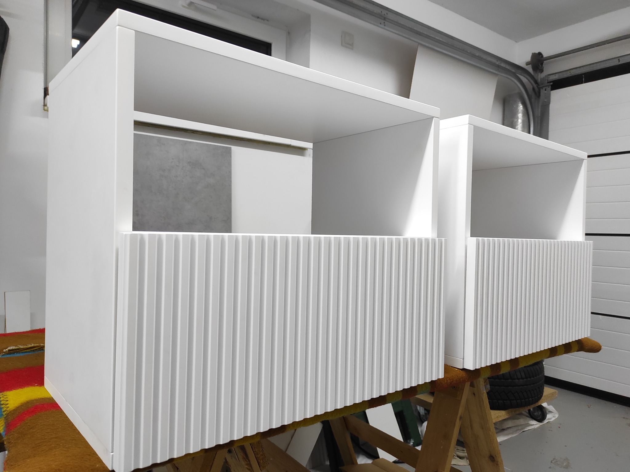 Białe, minimalistyczne szafki z ryflowanymi frontami, ustawione na drewnianych kozłach w warsztacie. Widoczny fragment opony i brama garażowa w tle.