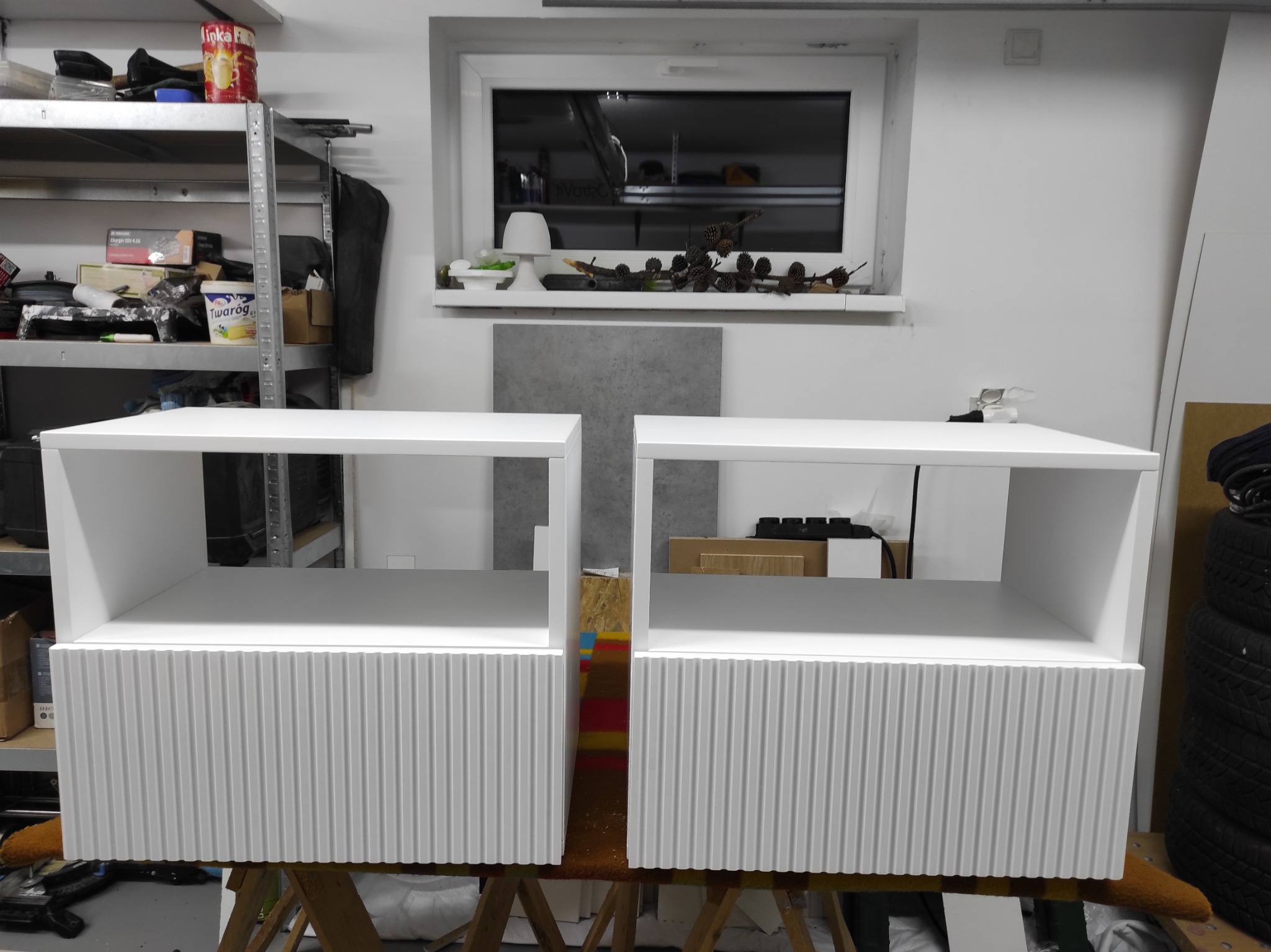 Dwie białe szafki nocne z ryflowanymi frontami, ustawione obok siebie w warsztacie, pod oknem z dekoracjami z szyszek. W tle regał z narzędziami.