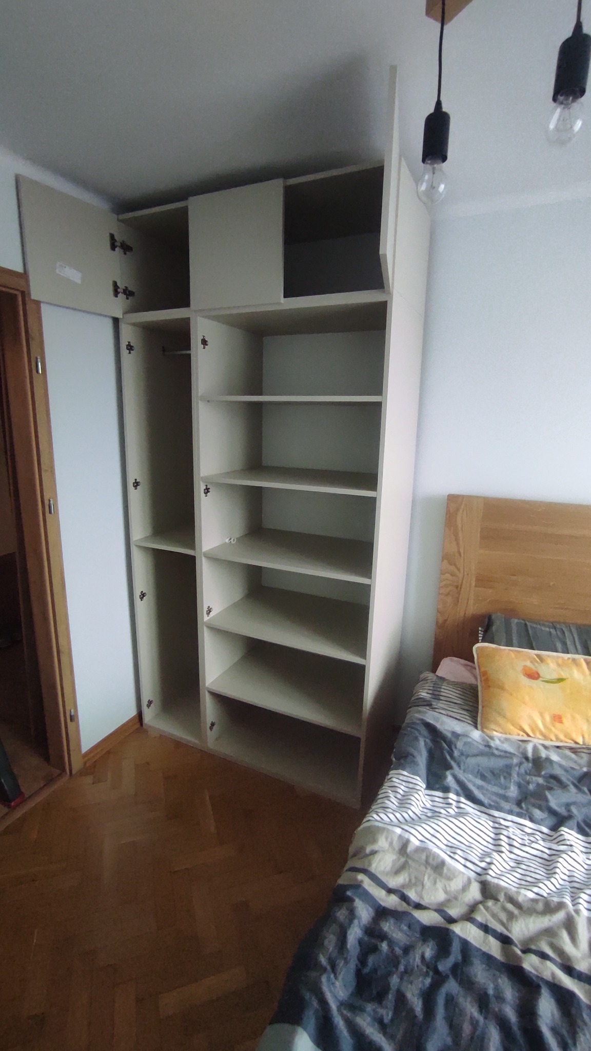 Jasna, modułowa szafa z otwartymi półkami i drzwiami, widoczna w sypialni w Rzeszowie. Parkiet na podłodze, łóżko obok szafy.