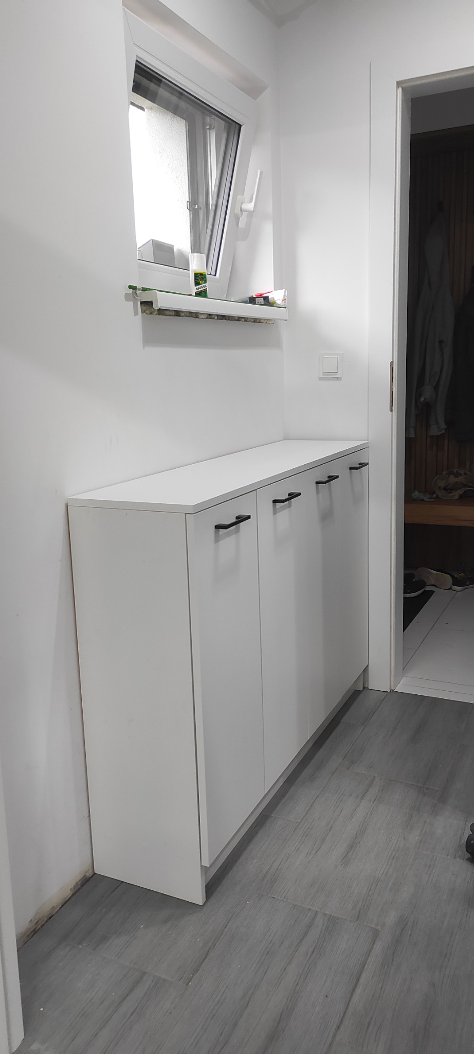 Biała szafka z trzema drzwiczkami i czarnymi uchwytami, stojąca w korytarzu na szarych panelach pod oknem. Widoczny fragment wnętrza szafy.