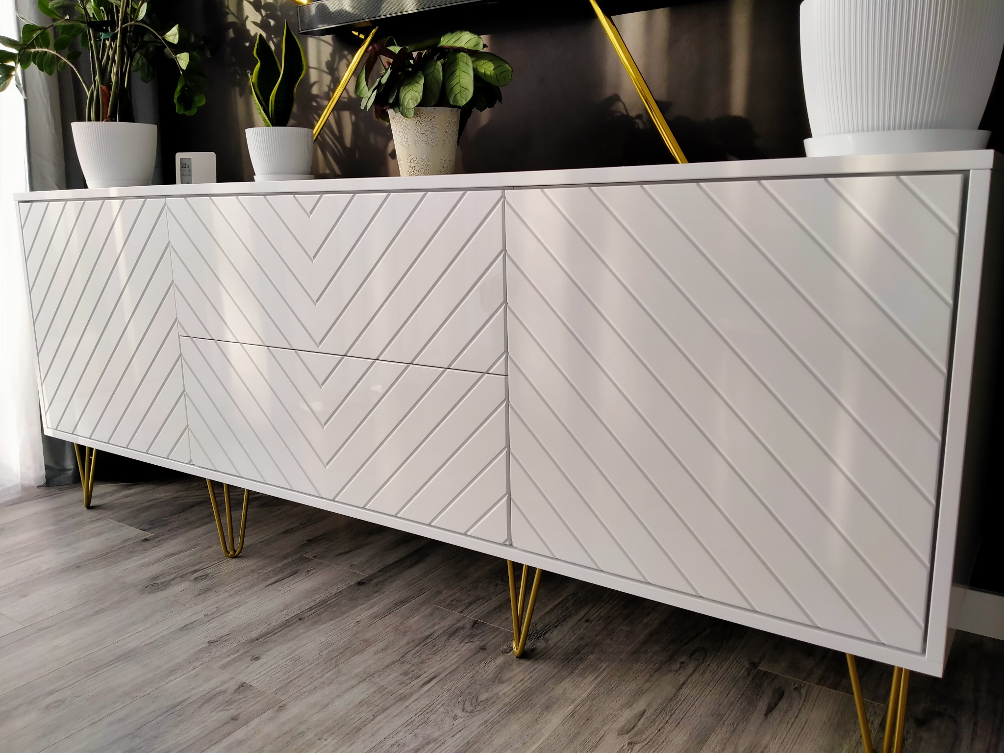 Biała komoda z geometrycznym wzorem jodełki na frontach, na złotych nóżkach. Na komodzie i za nią rośliny doniczkowe. Nowoczesny design.