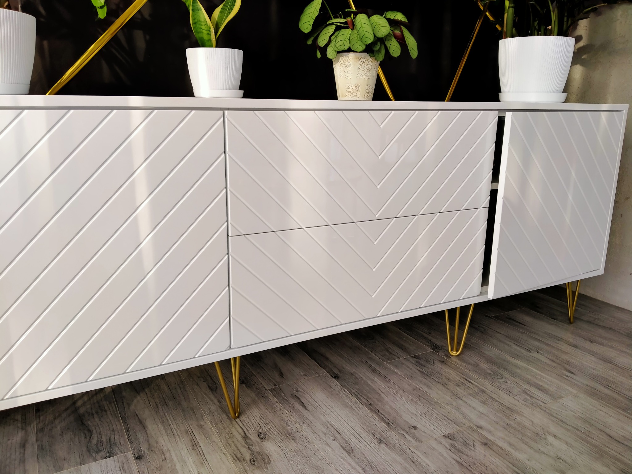 Biała komoda z frezowanym wzorem w jodełkę, na złotych nóżkach. Na blacie stoją doniczki z roślinami. Jedne drzwiczki są otwarte. Podłoga z paneli imitujących drewno.