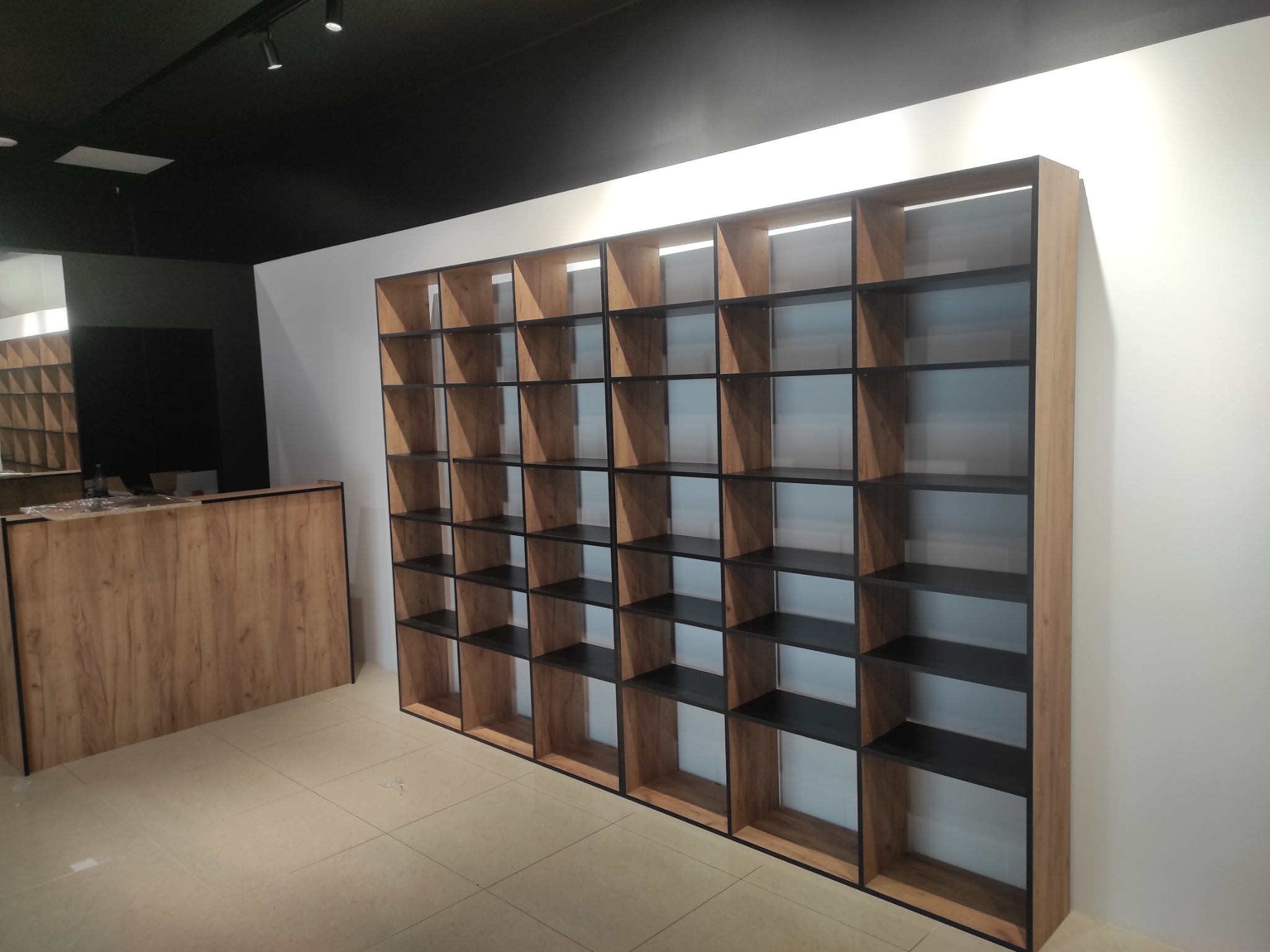 Regał z drewnianymi półkami o geometrycznym kształcie, z czarnymi wnętrzami, ustawiony przy białej ścianie w jasnym pomieszczeniu. Widoczny fragment lady.