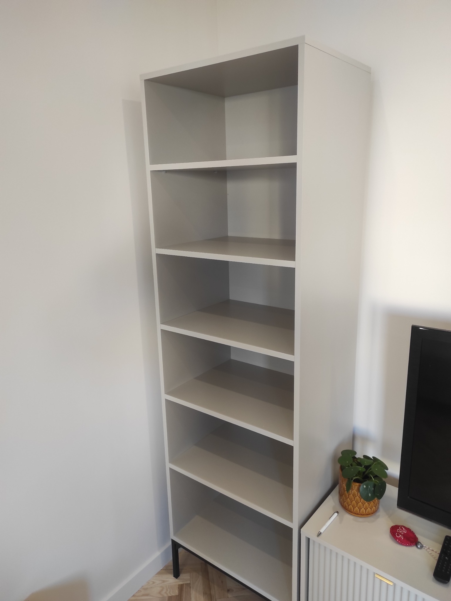 Jasnoszara, wysoka szafa regałowa z sześcioma półkami, ustawiona w rogu pokoju, z widocznymi czarnymi nóżkami i elementami dekoracyjnymi obok.