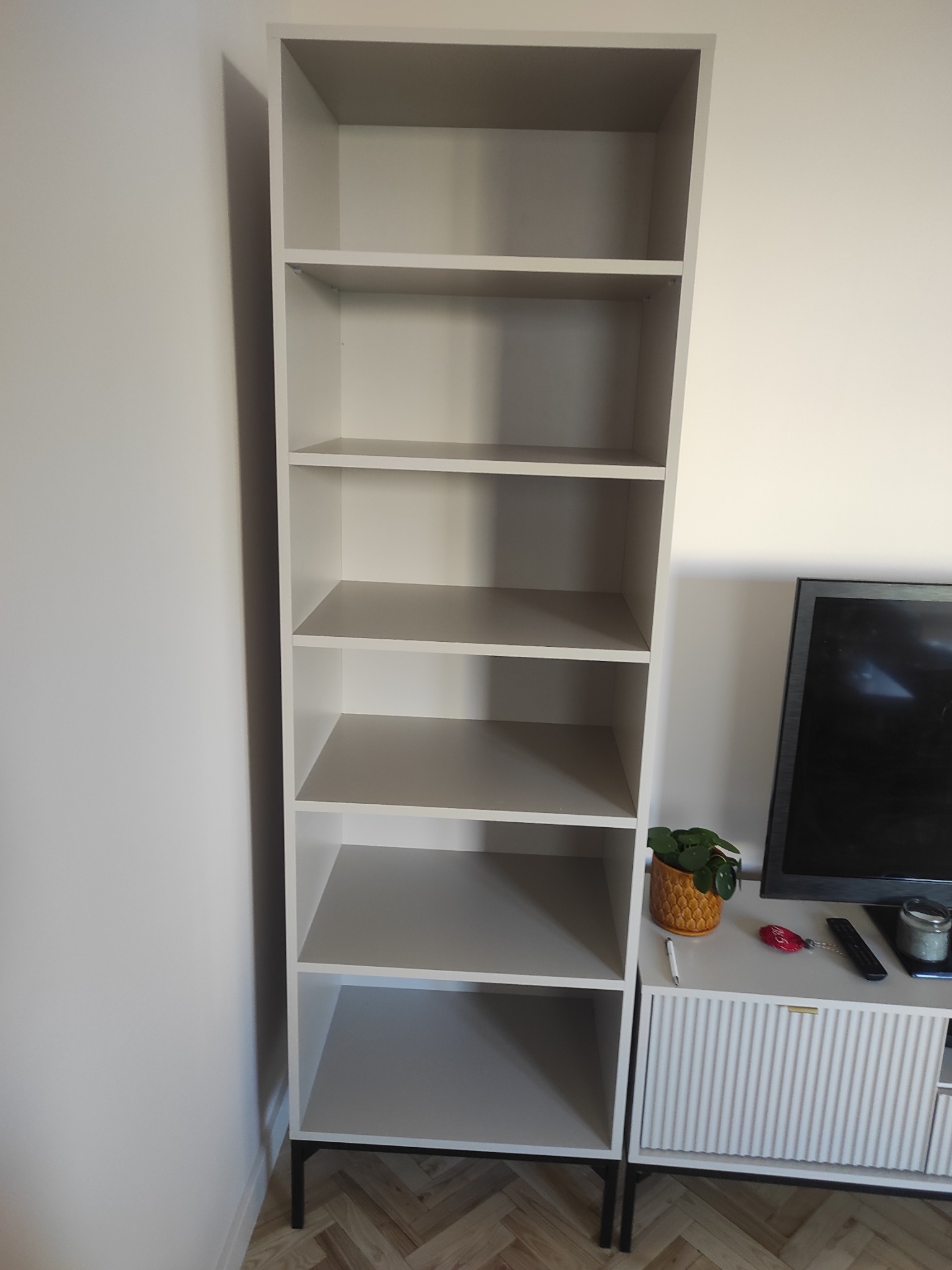 Wysoka, smukła, jasnoszara szafa regałowa z sześcioma półkami, na czarnych nóżkach, stojąca obok szafki RTV z telewizorem i dekoracjami.
