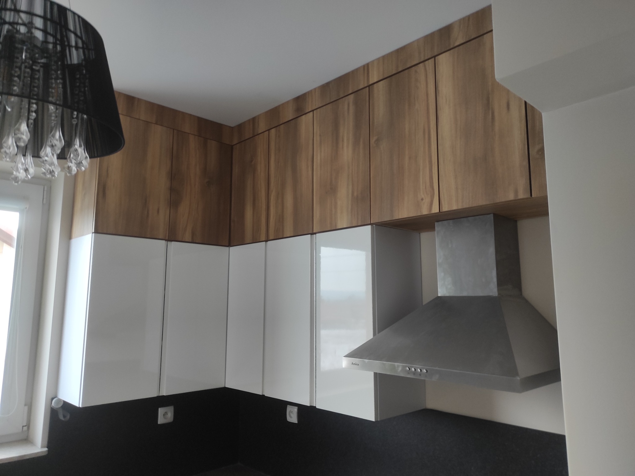 Nowoczesna kuchnia z białymi, błyszczącymi szafkami dolnymi i drewnianymi szafkami górnymi. Widoczny okap kuchenny i czarny blat. Uwagę przykuwa designerski żyrandol.