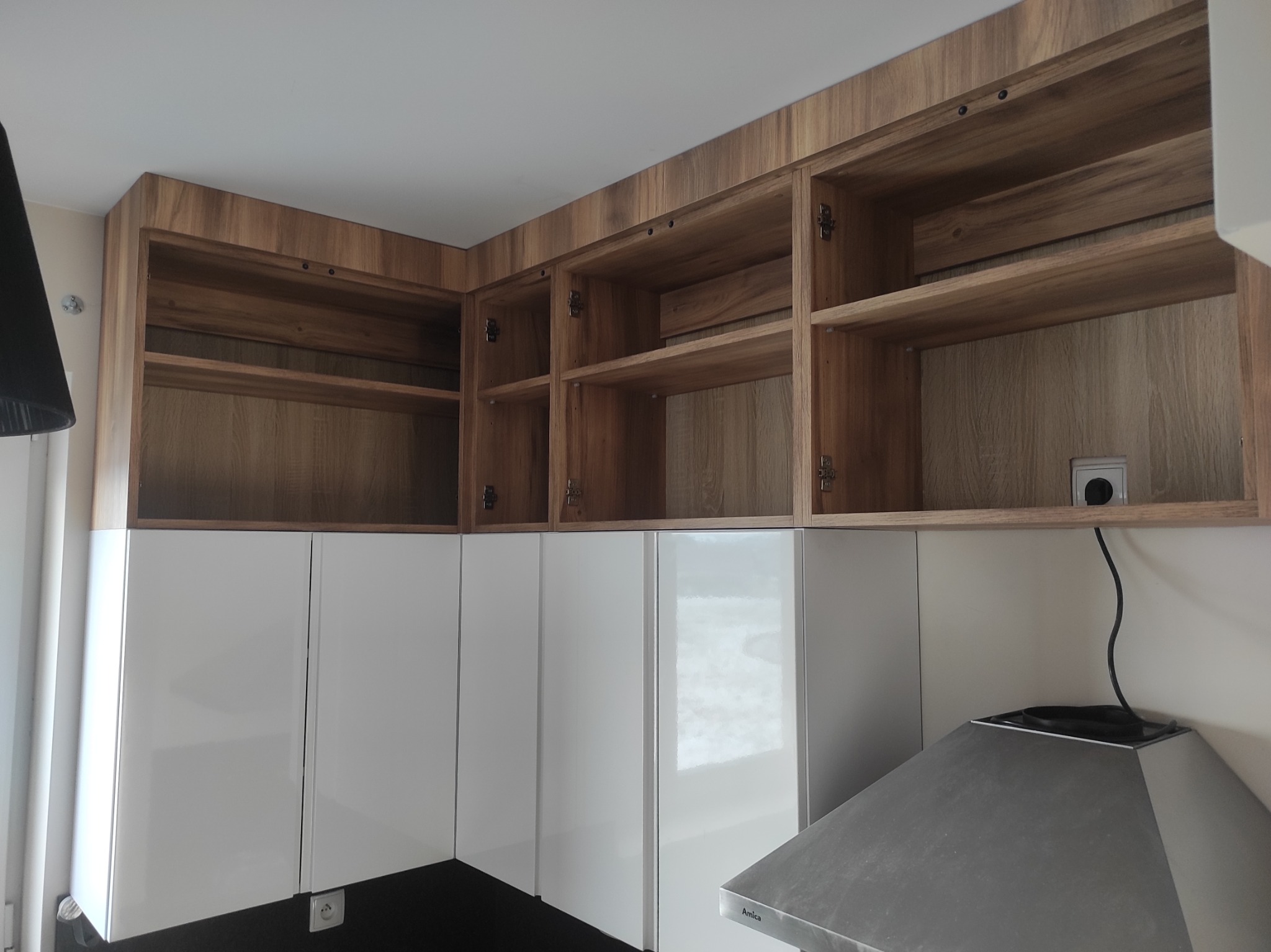 Nowoczesna kuchnia z białymi szafkami dolnymi i drewnianymi górnymi, otwartymi półkami. Widoczny okap kuchenny i gniazdko elektryczne na ścianie.