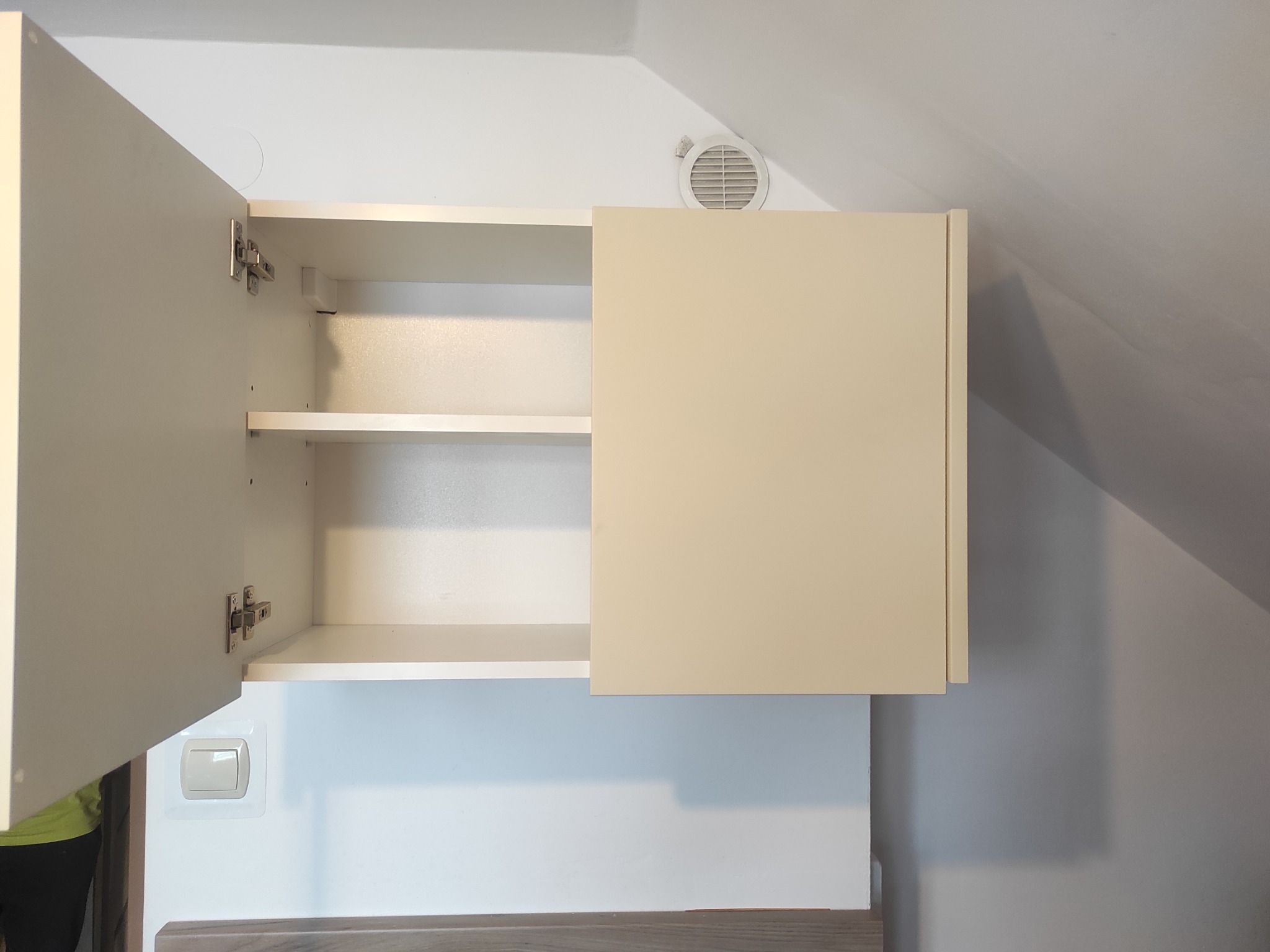 Biała szafka wisząca z otwartymi drzwiczkami, widoczne półki wewnątrz. Na ścianie kratka wentylacyjna i włącznik światła. Ujęcie z dołu, wnętrze w jasnych barwach.