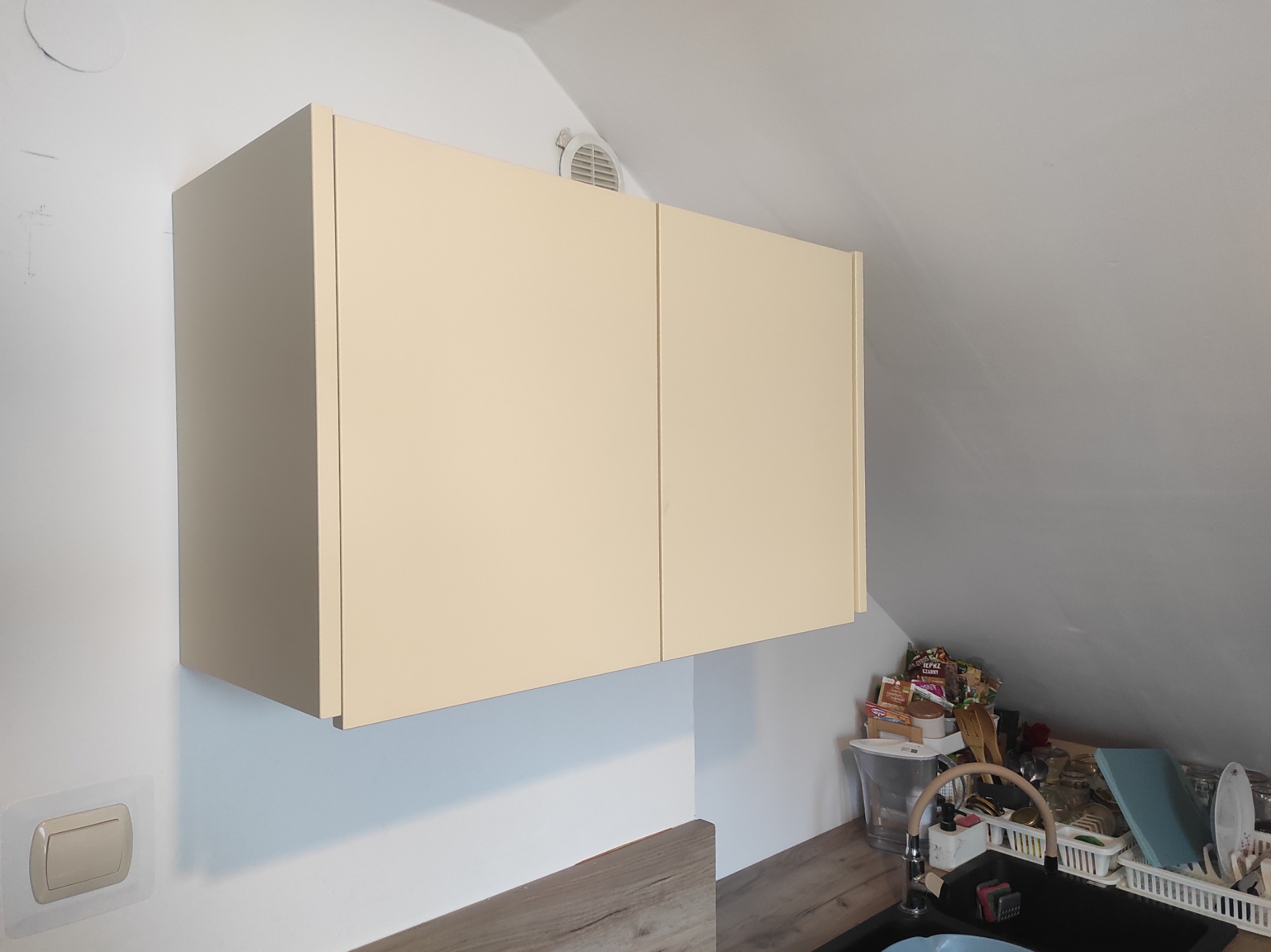 Beżowa szafka kuchenna pod skosem dachu, z widocznym fragmentem blatu i zlewu. Minimalistyczny design, jasne wnętrze, wentylacja w suficie.