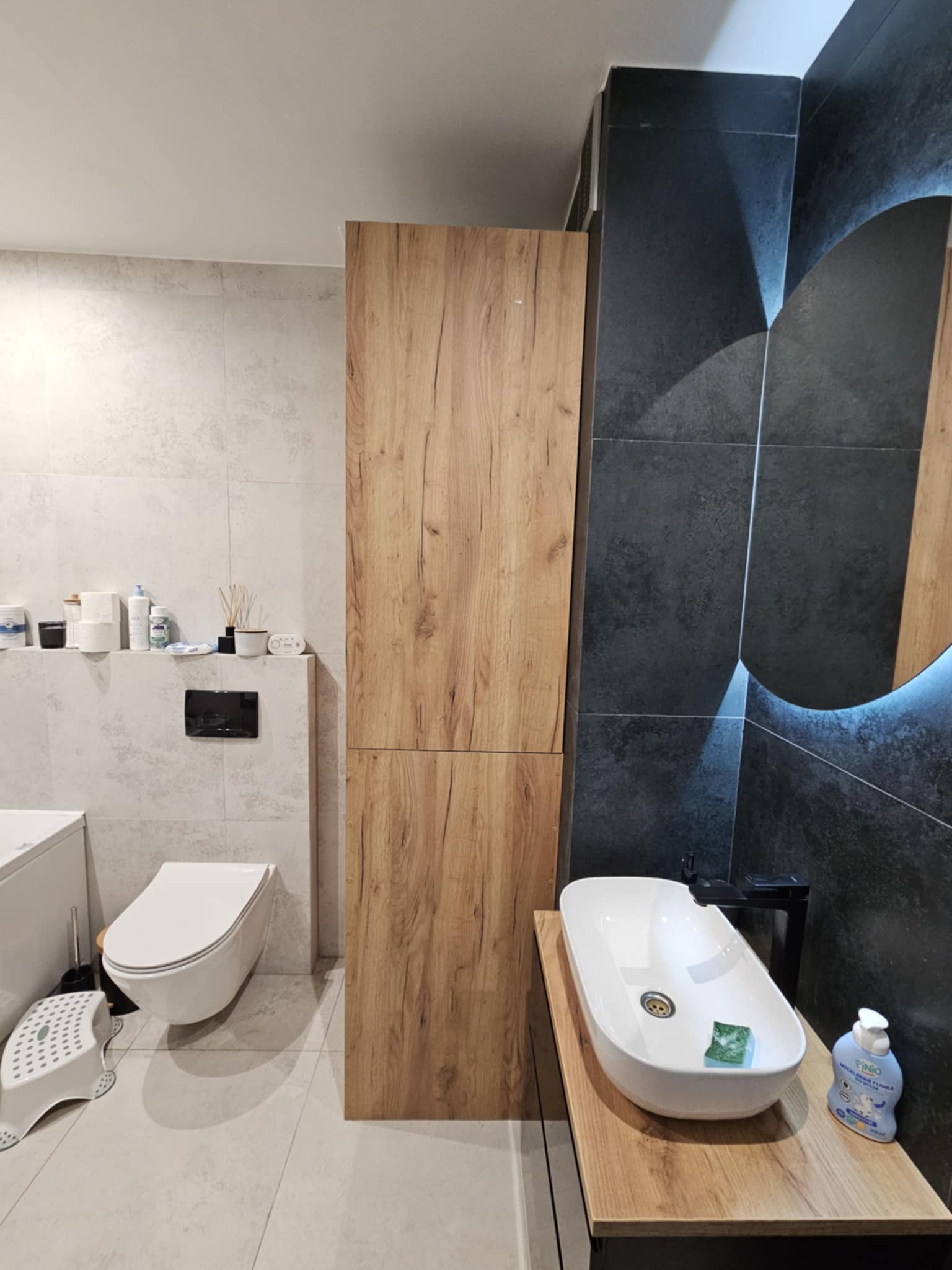 Nowoczesna łazienka z drewnianą szafką i blatem pod umywalkę, czarne płytki z podświetlanym lustrem, biała ceramika. Minimalistyczny design.