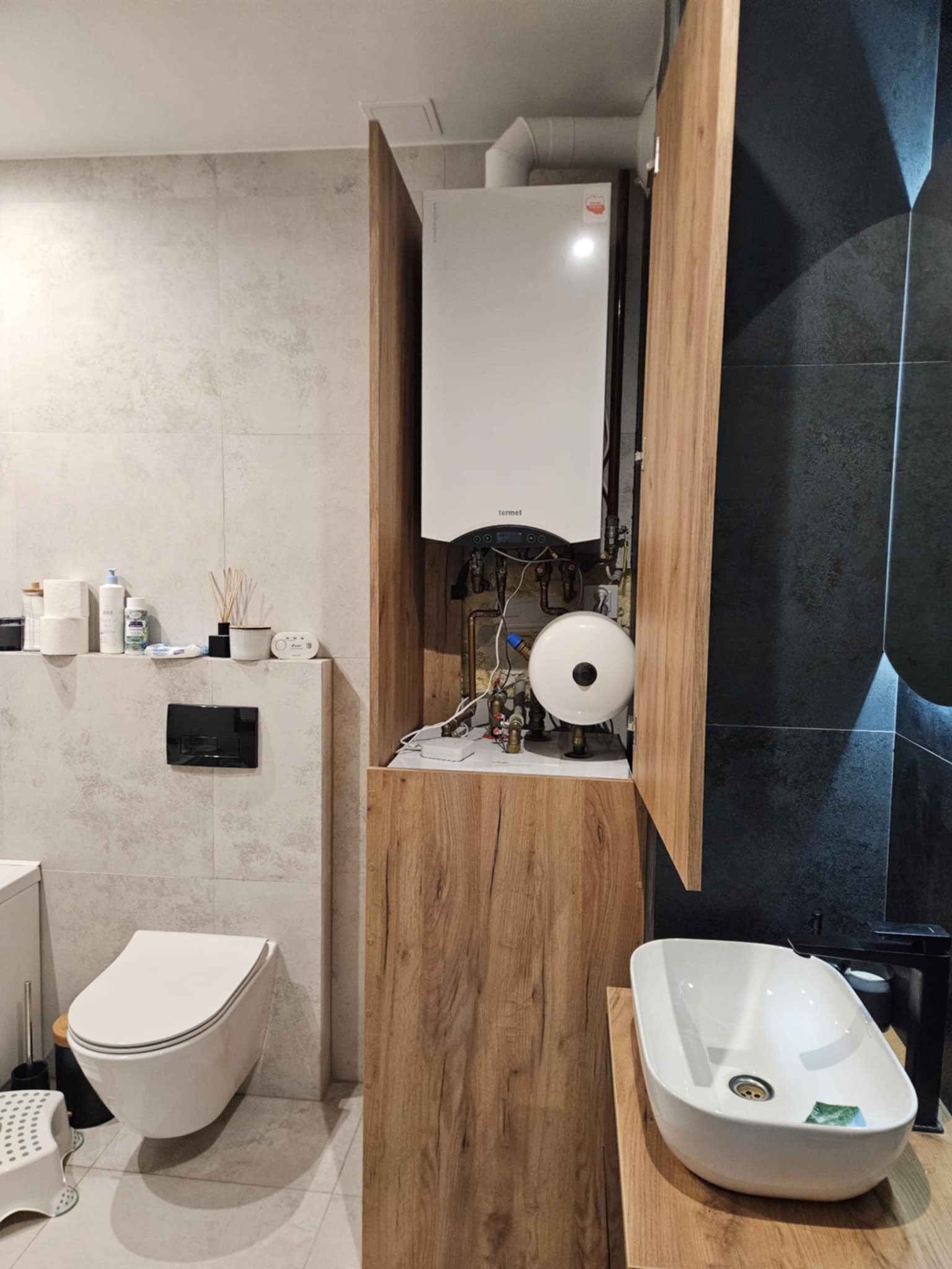 Nowoczesna łazienka z zabudowanym piecem gazowym w szafce z drewnianym frontem. Umywalka nablatowa i podwieszana toaleta. Szare i czarne płytki na ścianach.