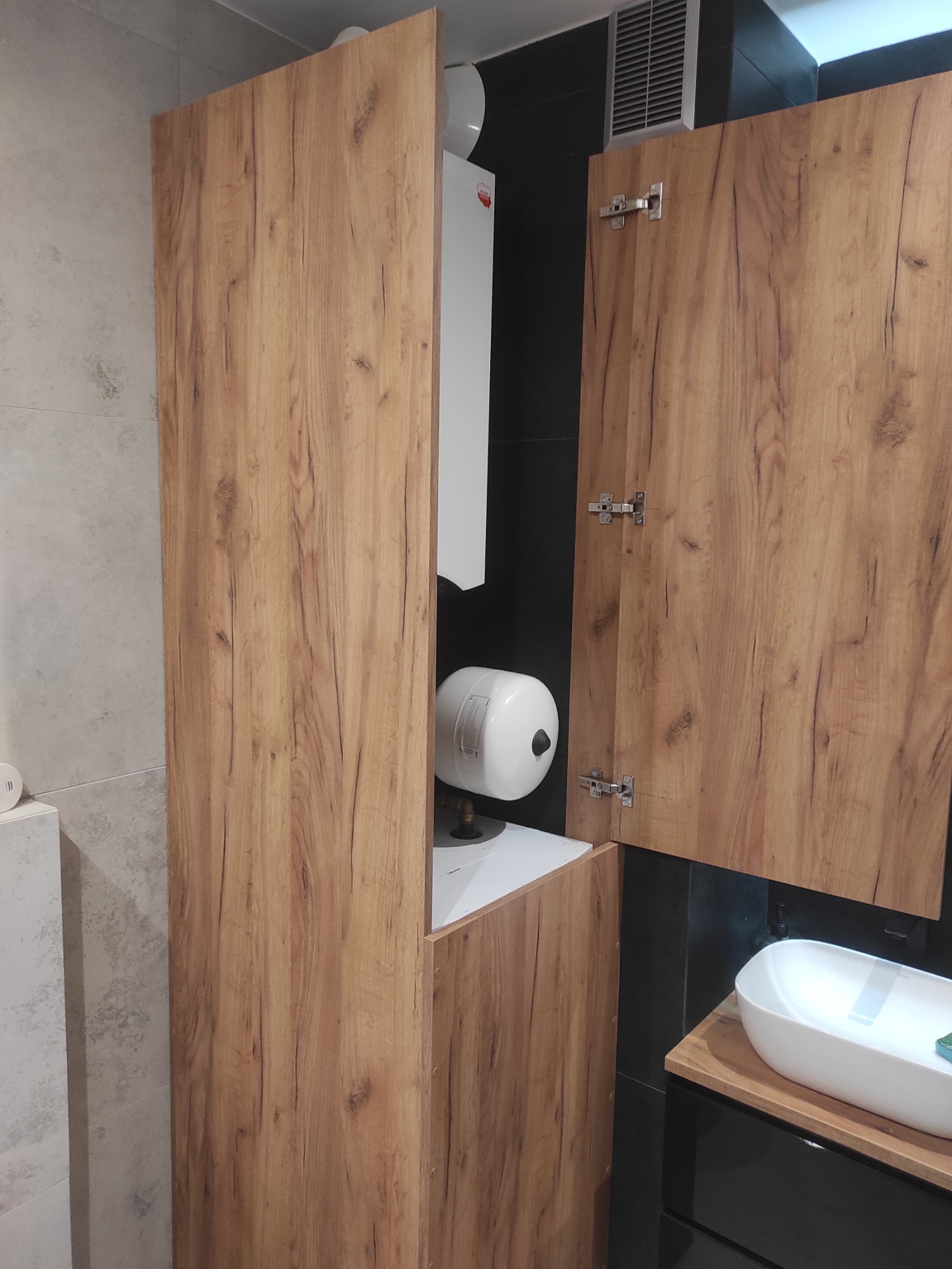 Szafka łazienkowa z drewnianymi frontami, częściowo otwarta, ukrywająca bojler. Widoczna umywalka nablatowa i czarne płytki w tle. Ujęcie z bliska.