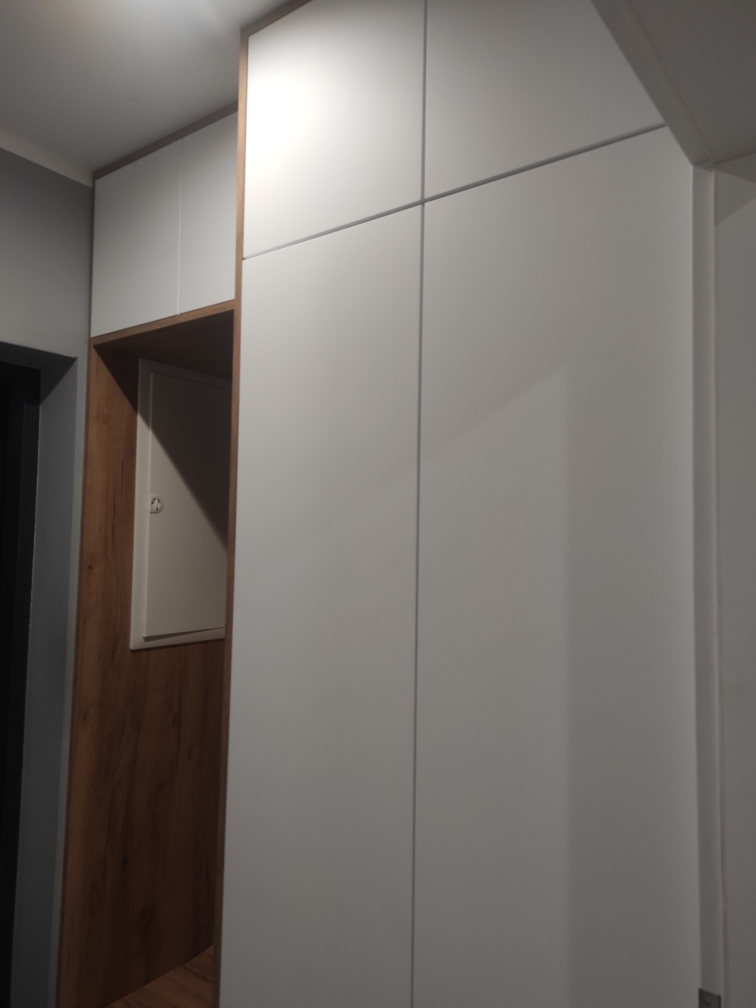 Biała szafa wnękowa z drewnianym akcentem i ukrytym panelem elektrycznym, minimalistyczny design, oświetlenie sufitowe.