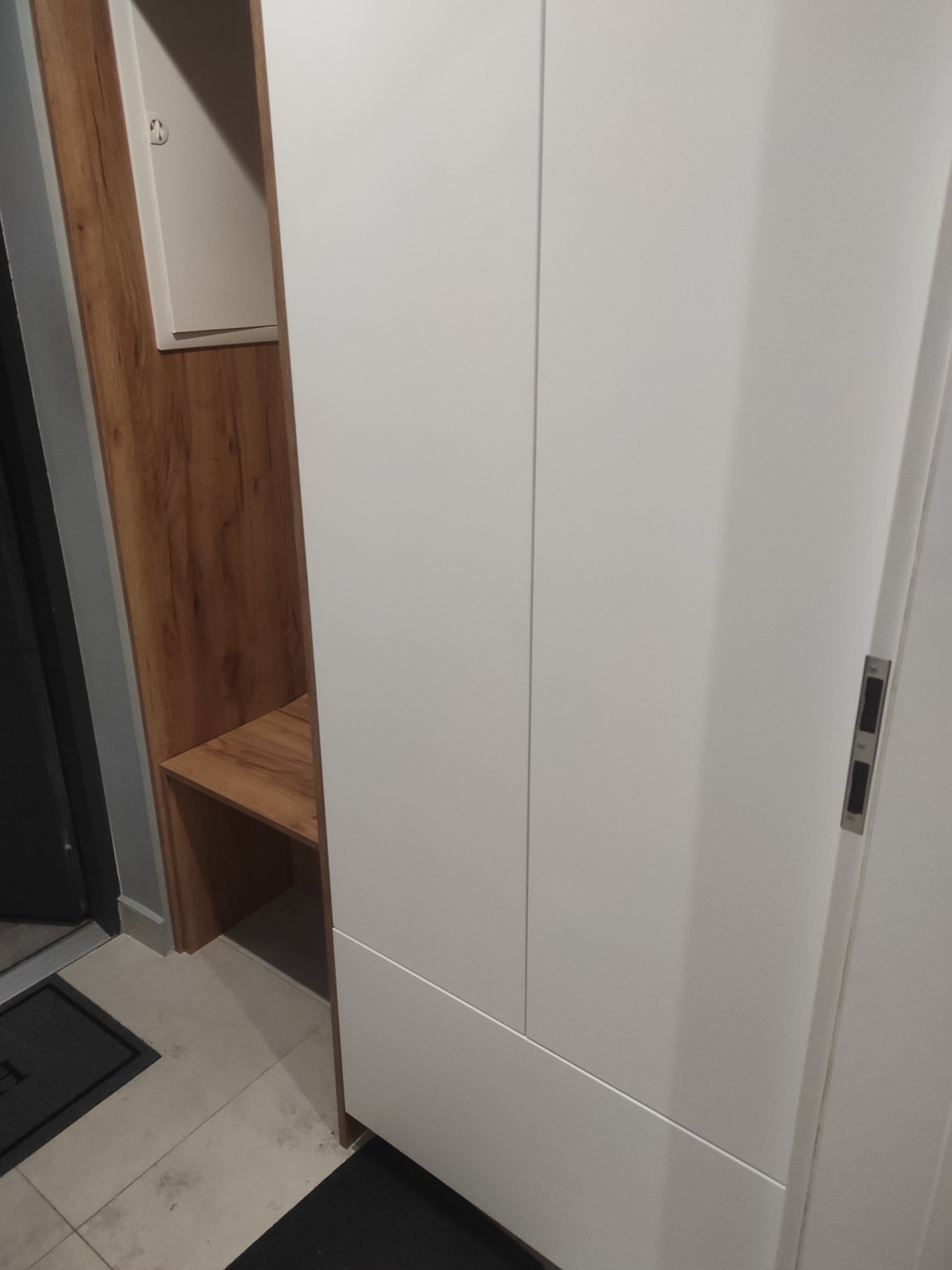 Biała szafa wnękowa z drewnianym akcentem i siedziskiem, widoczna przy wejściu do mieszkania. Minimalistyczny design, jasne kolory, nowoczesne wykończenie.