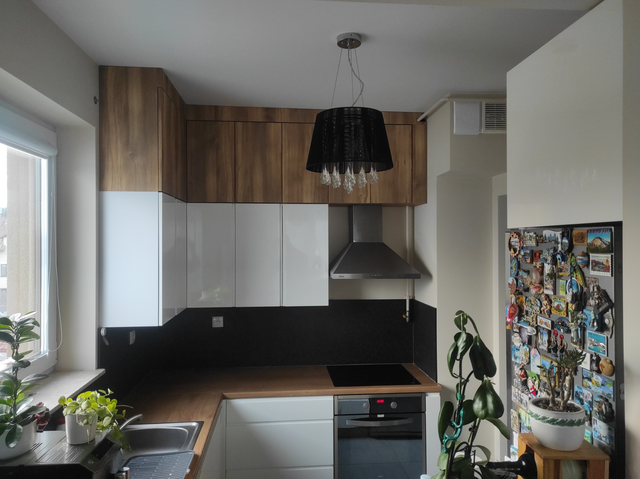 Nowoczesna kuchnia z białymi i drewnianymi szafkami, czarnym panelem nad blatem i designerską lampą. Ujęcie z perspektywy użytkownika, widoczne rośliny doniczkowe.