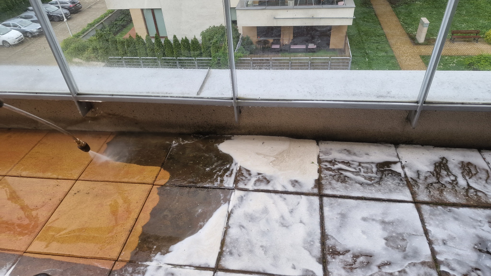 Czyszczenie płytek na balkonie za pomocą myjki ciśnieniowej. Widoczne efekty czyszczenia i piana. Tło stanowi widok na budynek i zieleń.