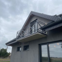 Przemysław Bąk - Nowoczesny balkon z geometryczną, czarną balustradą metalową, widoczny z dołu na tle pochmurnego nieba. Minimalistyczny design, szara elewacja budynku.