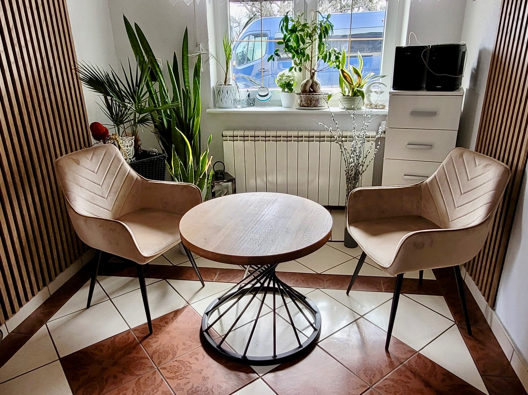 Stylowe wnętrze z okrągłym stolikiem kawowym na metalowej podstawie, dwoma krzesłami z tapicerką w odcieniach beżu i roślinami doniczkowymi przy oknie.