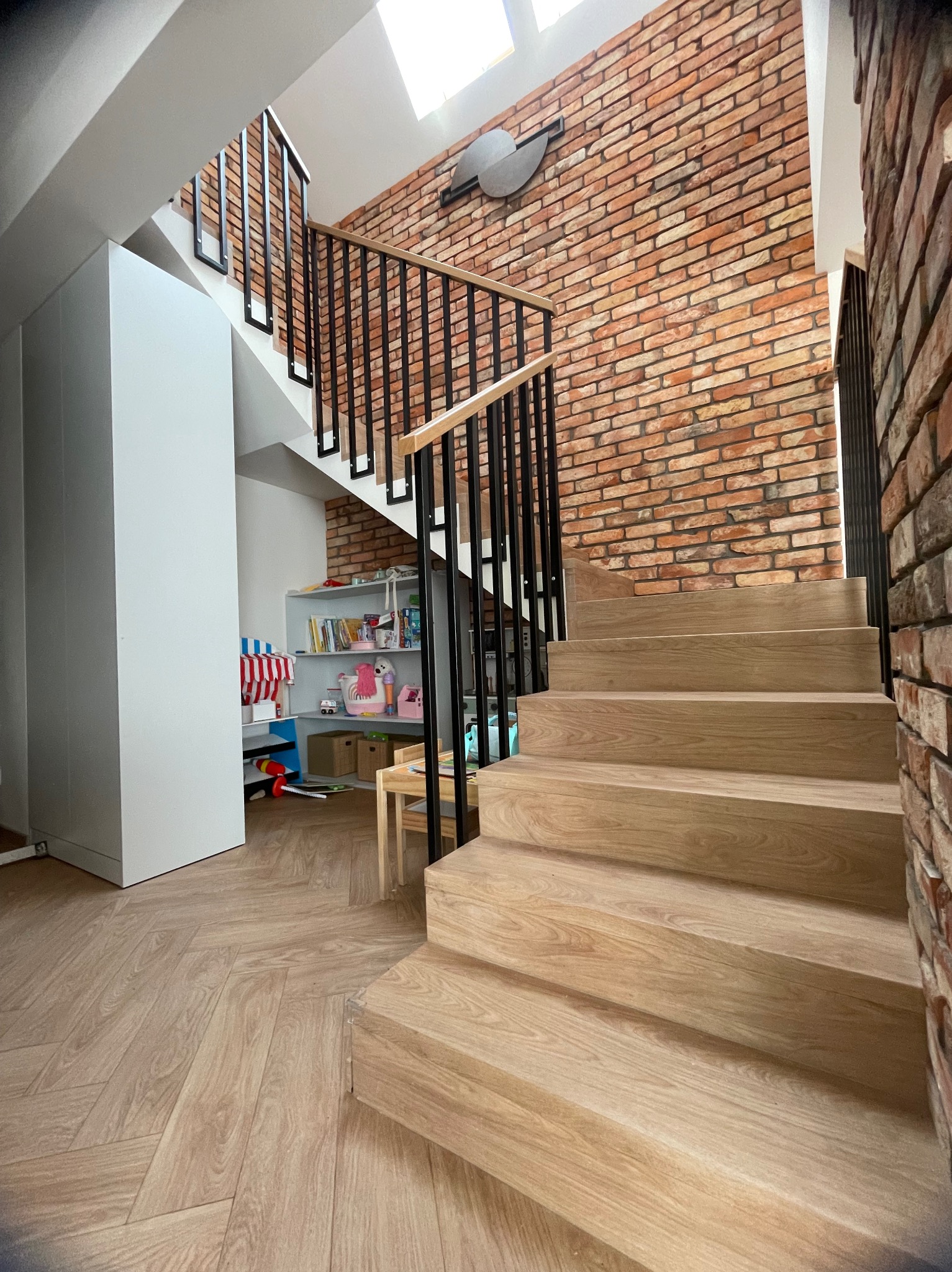Drewniane schody z czarną, metalową balustradą w nowoczesnym wnętrzu z ceglaną ścianą i jasną podłogą. Widok z dołu, podkreślający solidność konstrukcji.