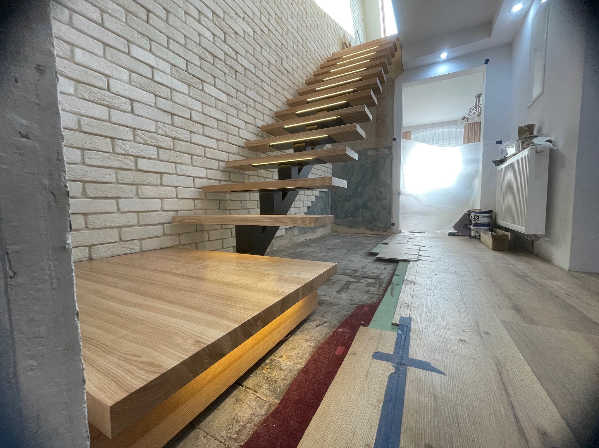 Nowoczesne schody drewniane z podświetleniem LED, wspornikowe, zamontowane przy ścianie z białej cegły. Widoczny fragment podłogi w trakcie montażu.