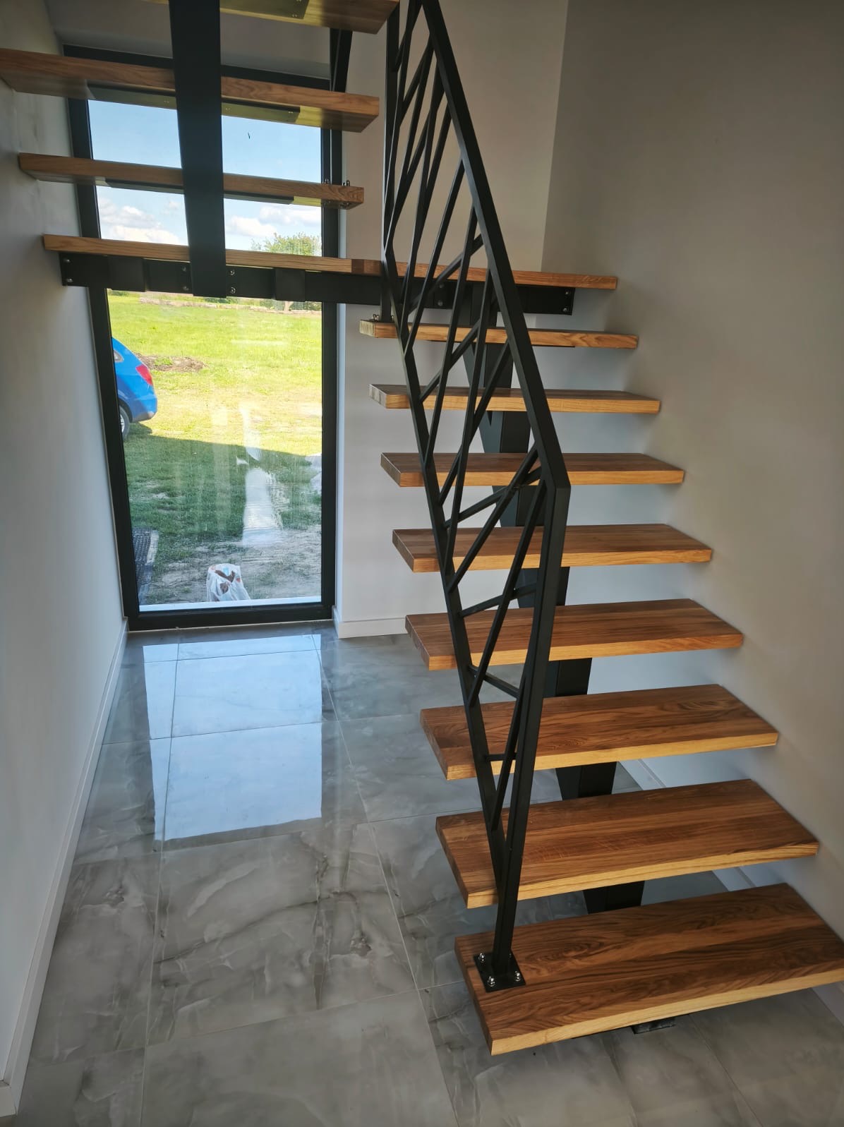 Nowoczesne schody z drewnianymi stopniami i czarną, geometryczną balustradą metalową. Widok na zewnątrz przez okno. Szare płytki podłogowe.
