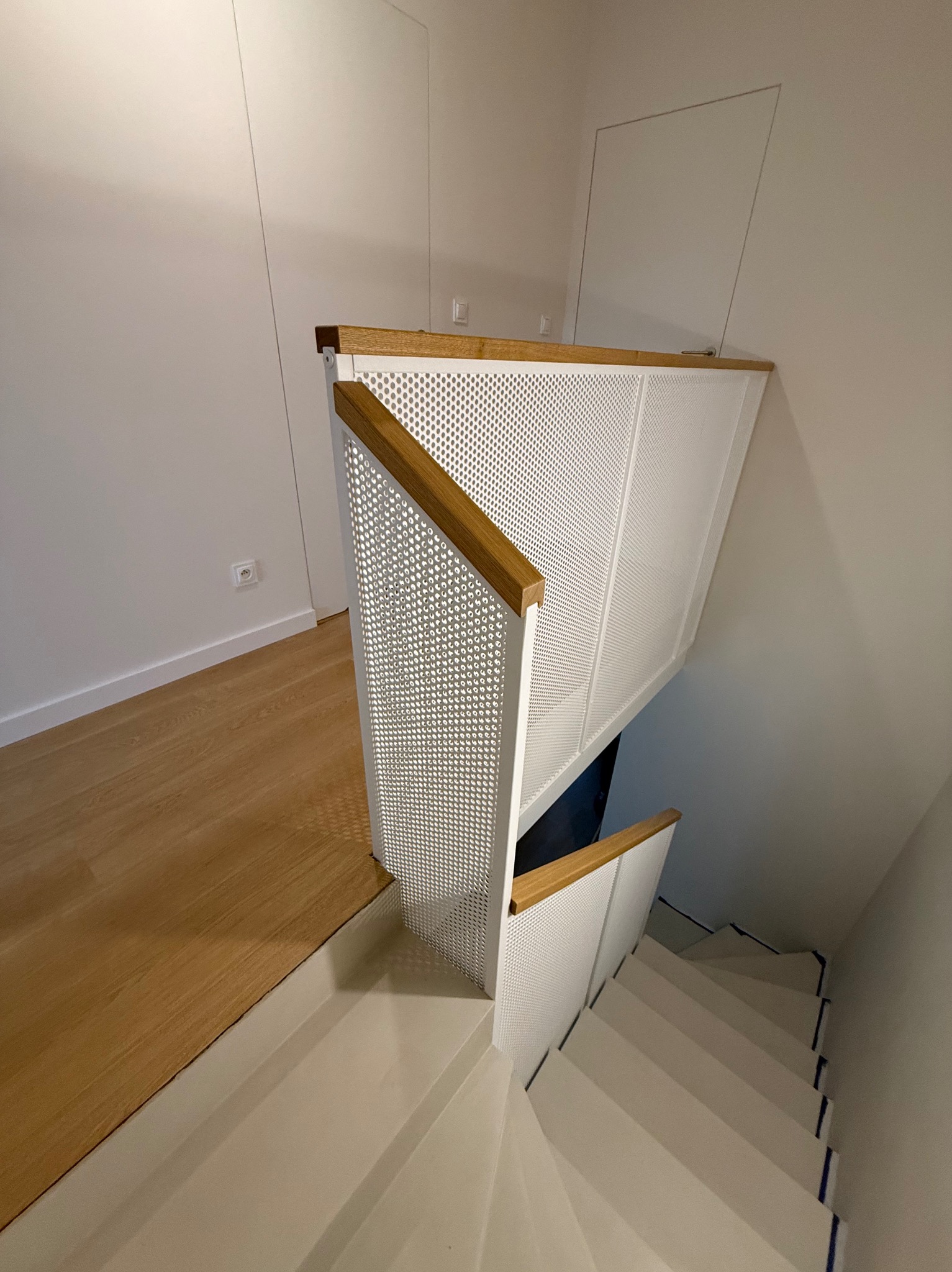 Nowoczesne schody z białą, perforowaną balustradą i drewnianym poręczem, widziane z góry. Minimalistyczny design, jasne wnętrze, drewniana podłoga.