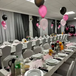 Uroczysty stół z białymi nakryciami, różową dekoracją i balonami z liczbą 18, przygotowany na przyjęcie urodzinowe w jasnej sali.