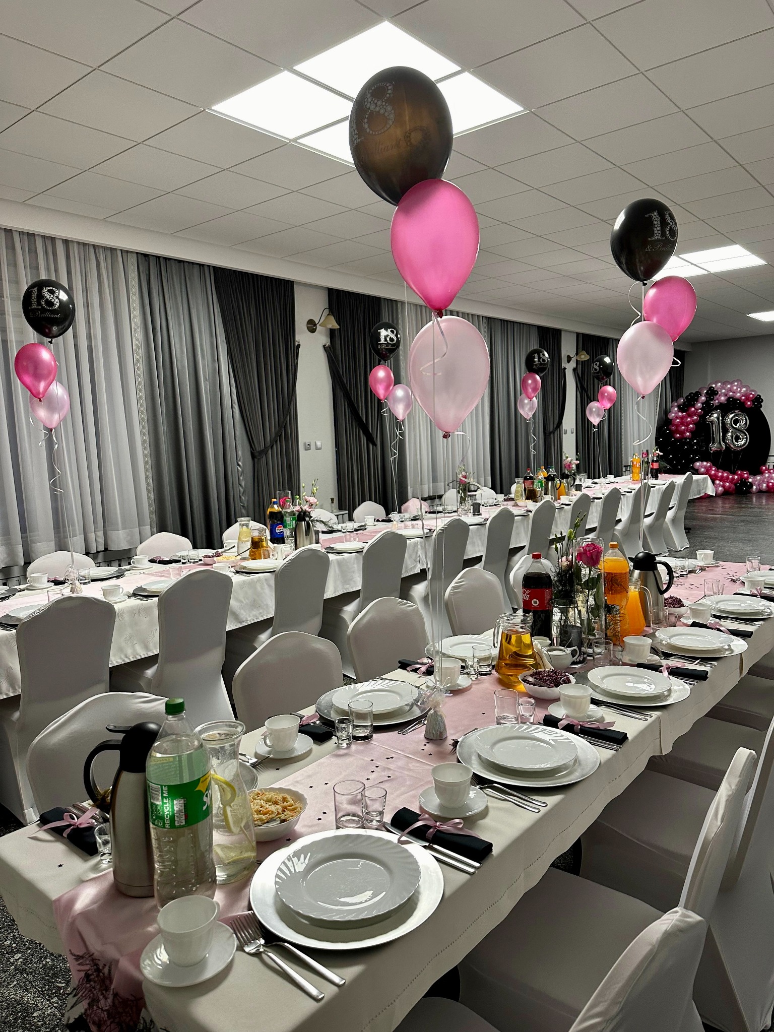 Uroczysty stół z białymi nakryciami, różową dekoracją i balonami z liczbą 18, przygotowany na przyjęcie urodzinowe w jasnej sali.