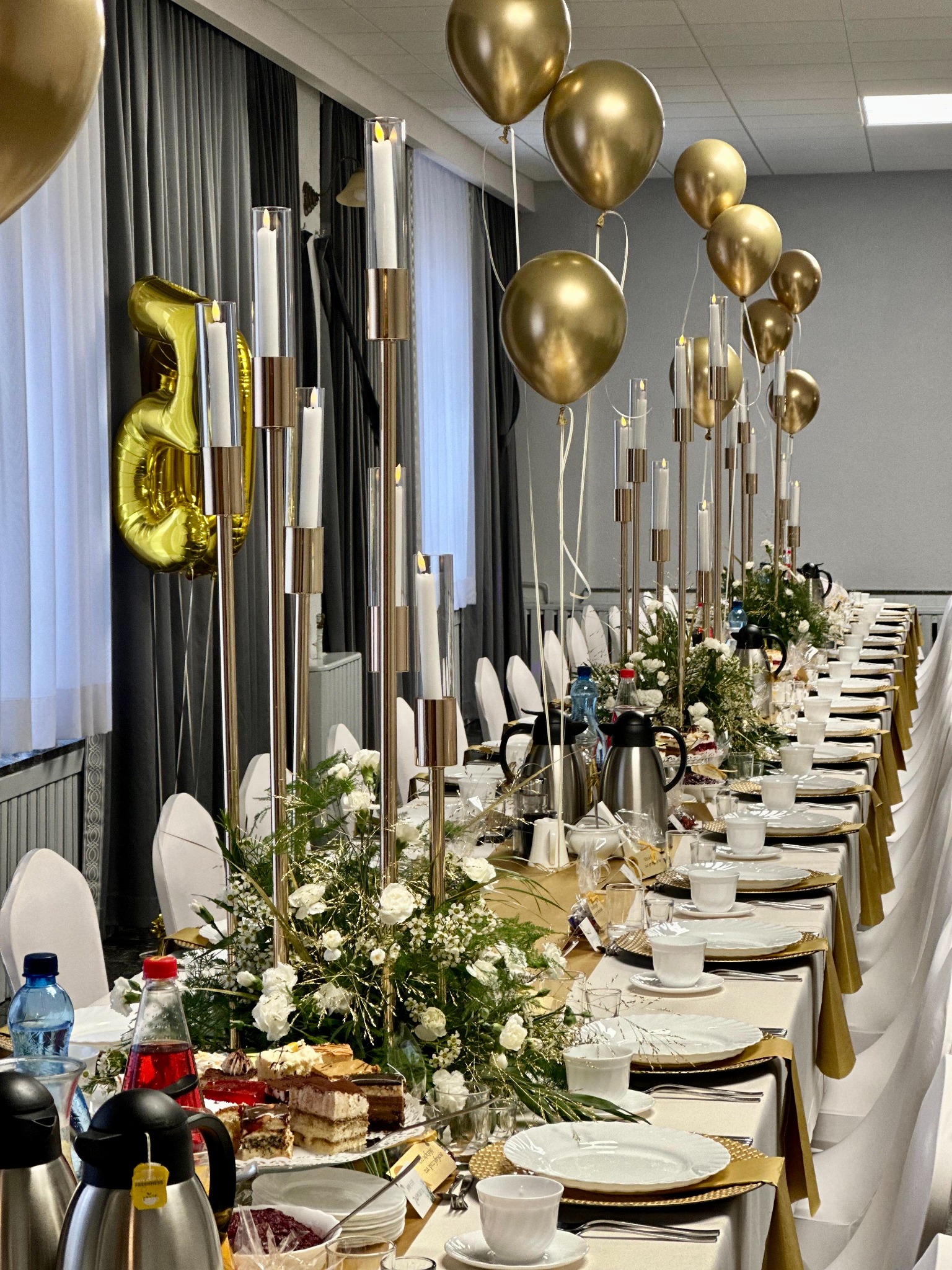 Uroczysty stół z białymi nakryciami, złotymi podkładkami, świecznikami i balonami. Widoczne napoje, ciasta i dekoracje kwiatowe. Przygotowania do przyjęcia.