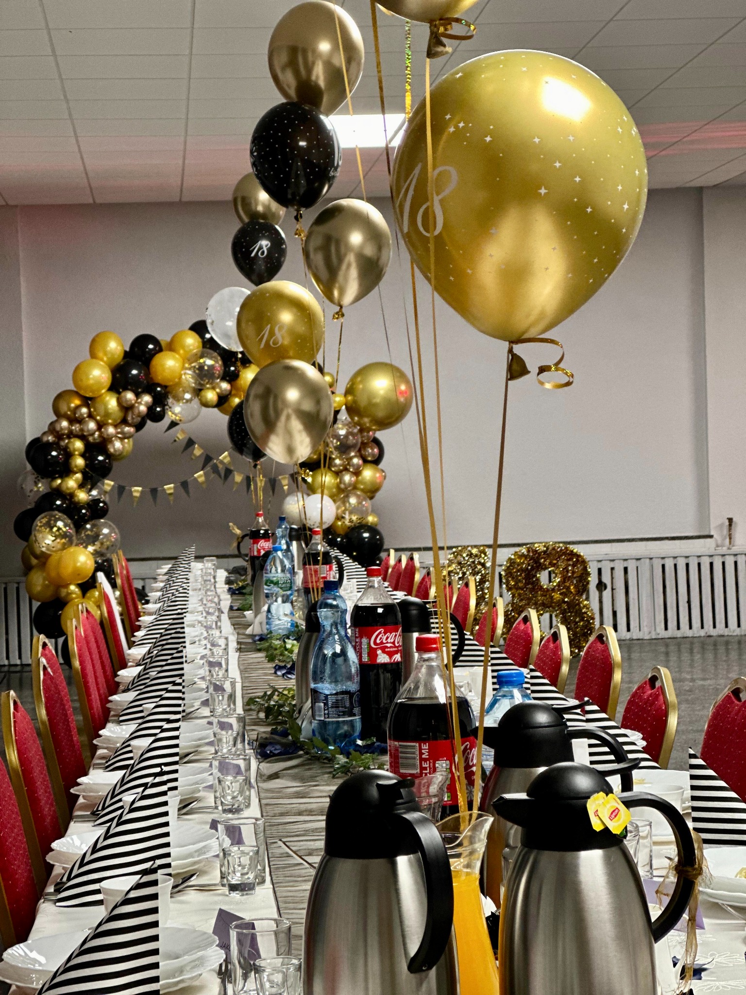 Uroczysty stół na 18 urodziny z dekoracją balonową w odcieniach złota i czerni, napojami, zastawą i czerwonymi krzesłami. Dekoracje urodzinowe.
