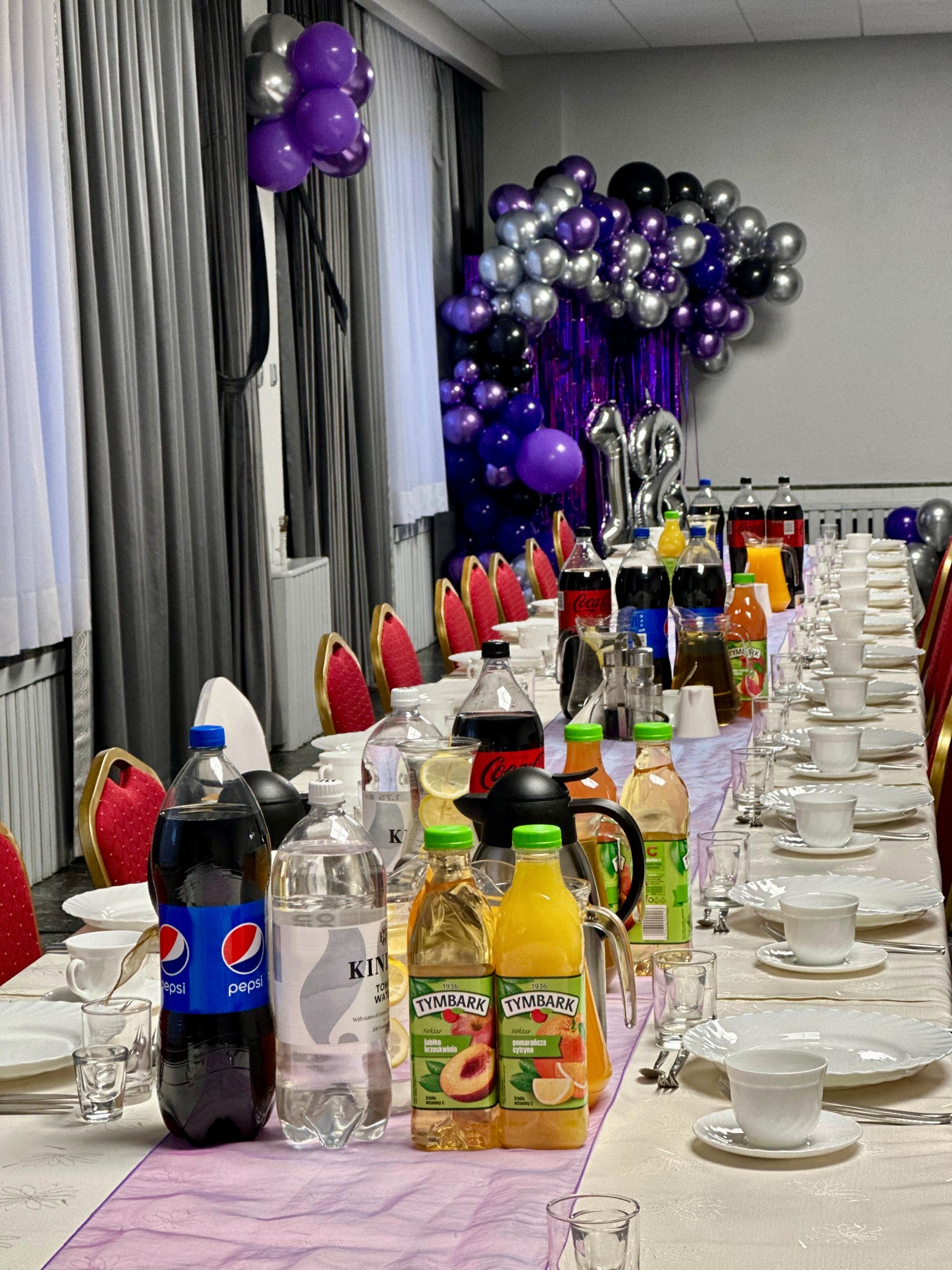 Długi stół z białym obrusem, nakryty zastawą i napojami. Dekoracje z balonów w odcieniach fioletu i srebra, tworzą klimat imprezy okolicznościowej.
