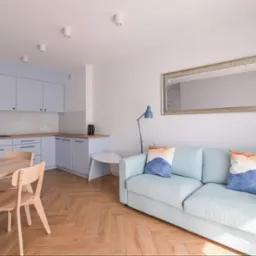 Jasne, przestronne wnętrze po remoncie: drewniana podłoga w jodełkę, błękitne meble kuchenne i wypoczynkowe, okrągły stół z krzesłami, lustro w ozdobnej ramie.