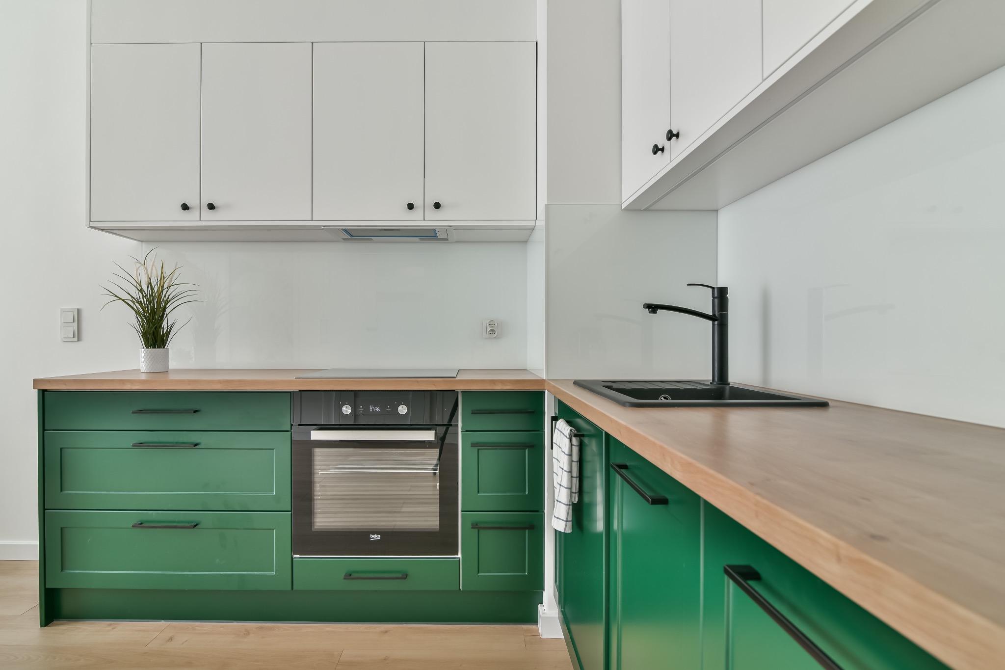 Nowoczesna kuchnia z zielonymi szafkami, drewnianym blatem i białymi szafkami wiszącymi. Minimalistyczny design z czarną armaturą i sprzętem AGD.